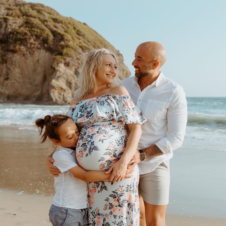 A happy family with a pregnant mother, embracing child, and smiling father standing on the beach.