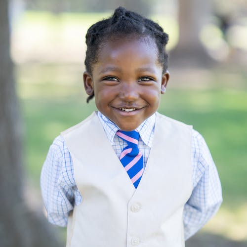 A young child in a vest and tie smiling in a park setting.
