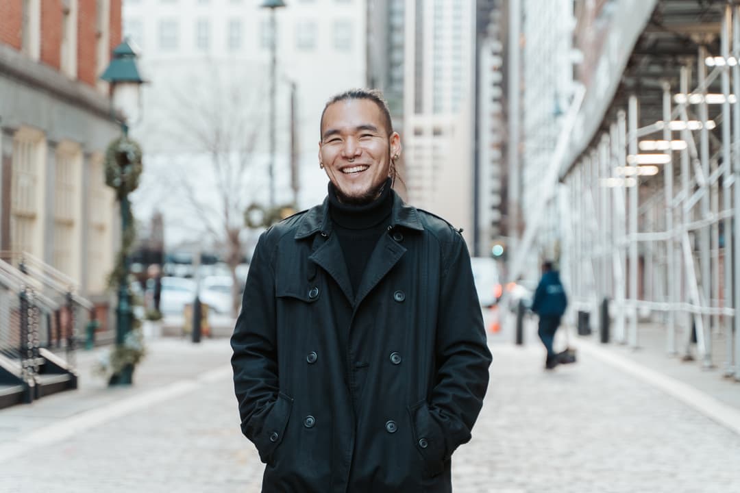 A smiling man with long hair wearing a black coat and turtleneck on an urban street.