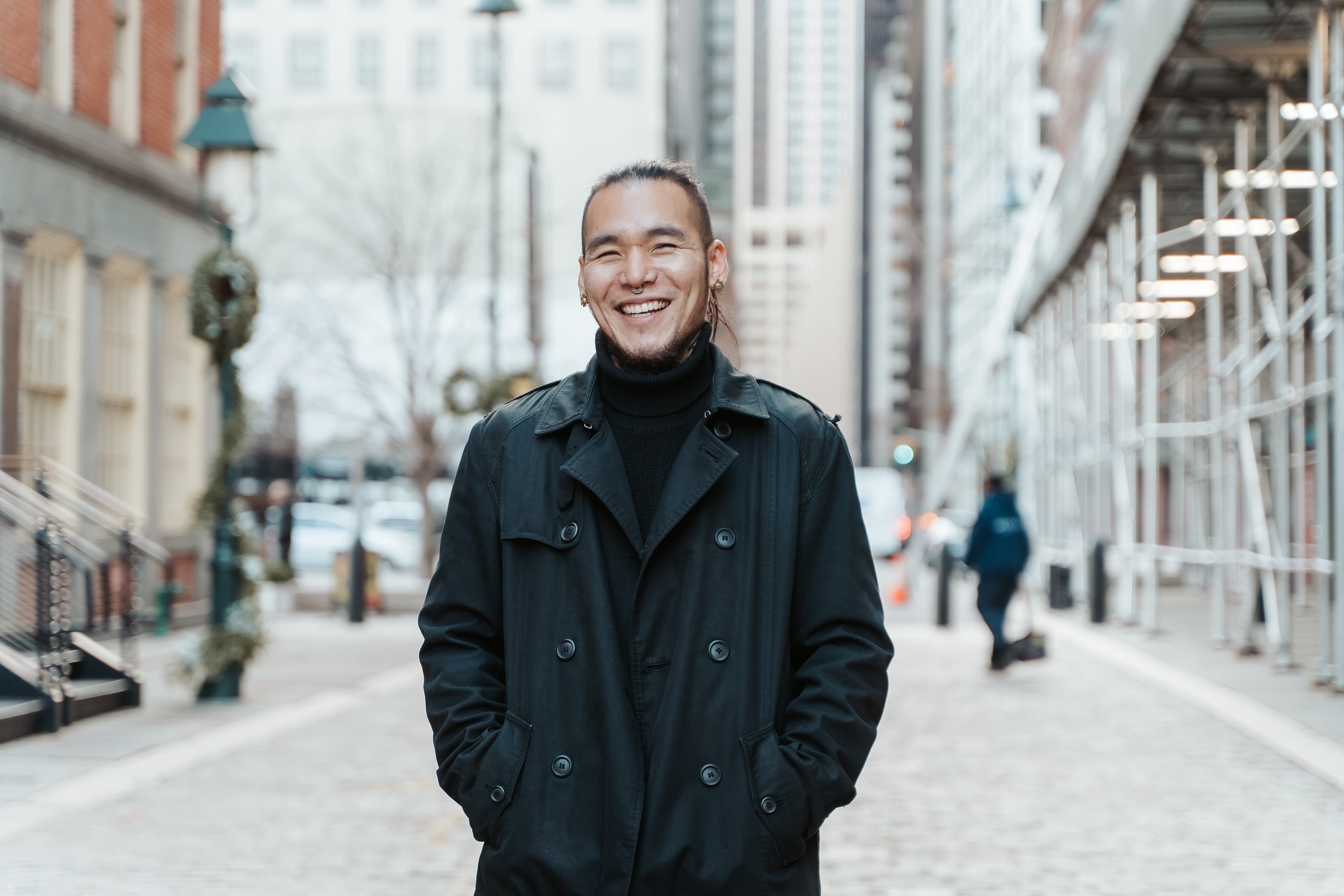 A smiling man with long hair wearing a black coat and turtleneck on an urban street.