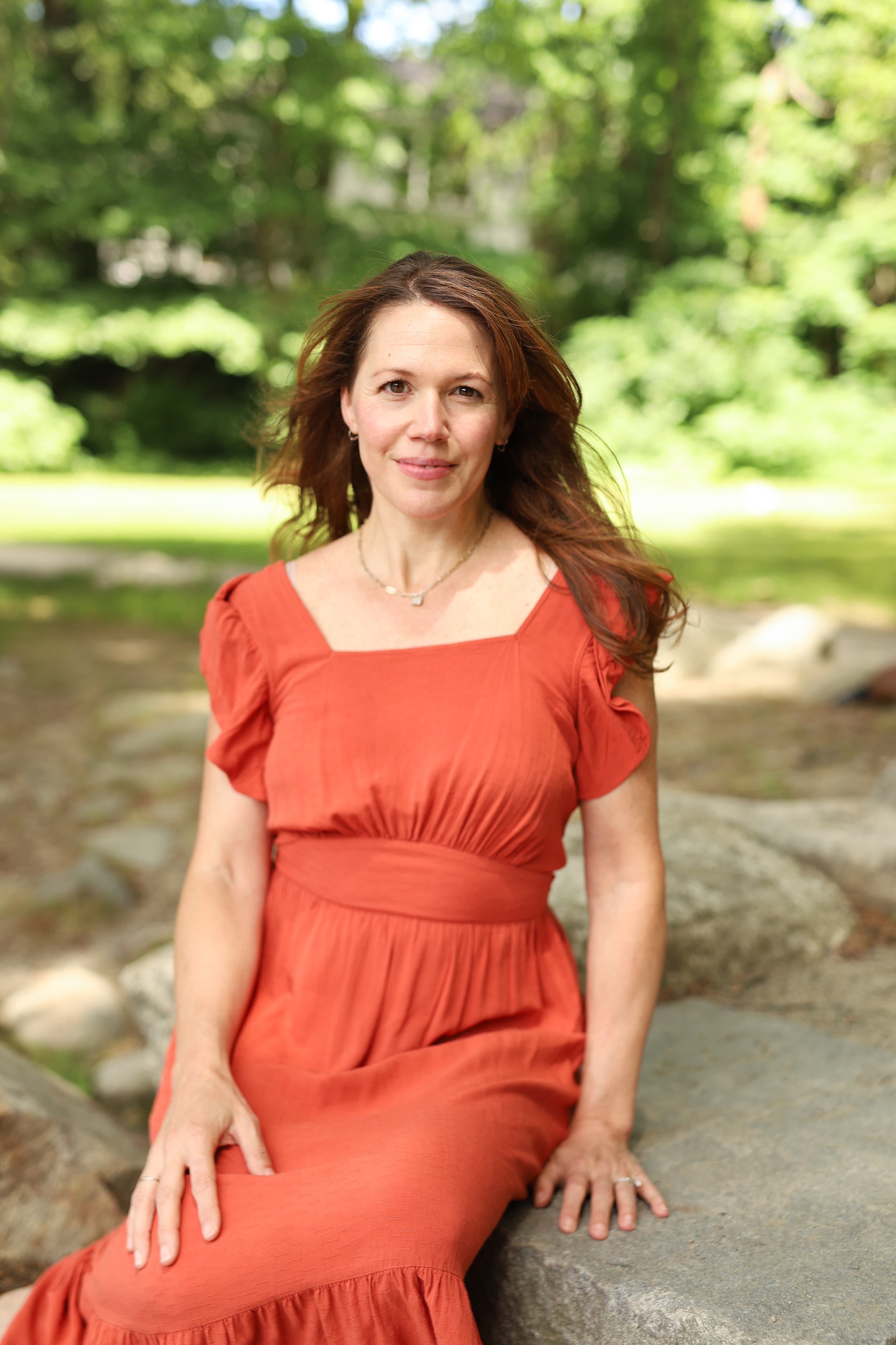 Woman in a red dress sitting on a rock with trees in the background.