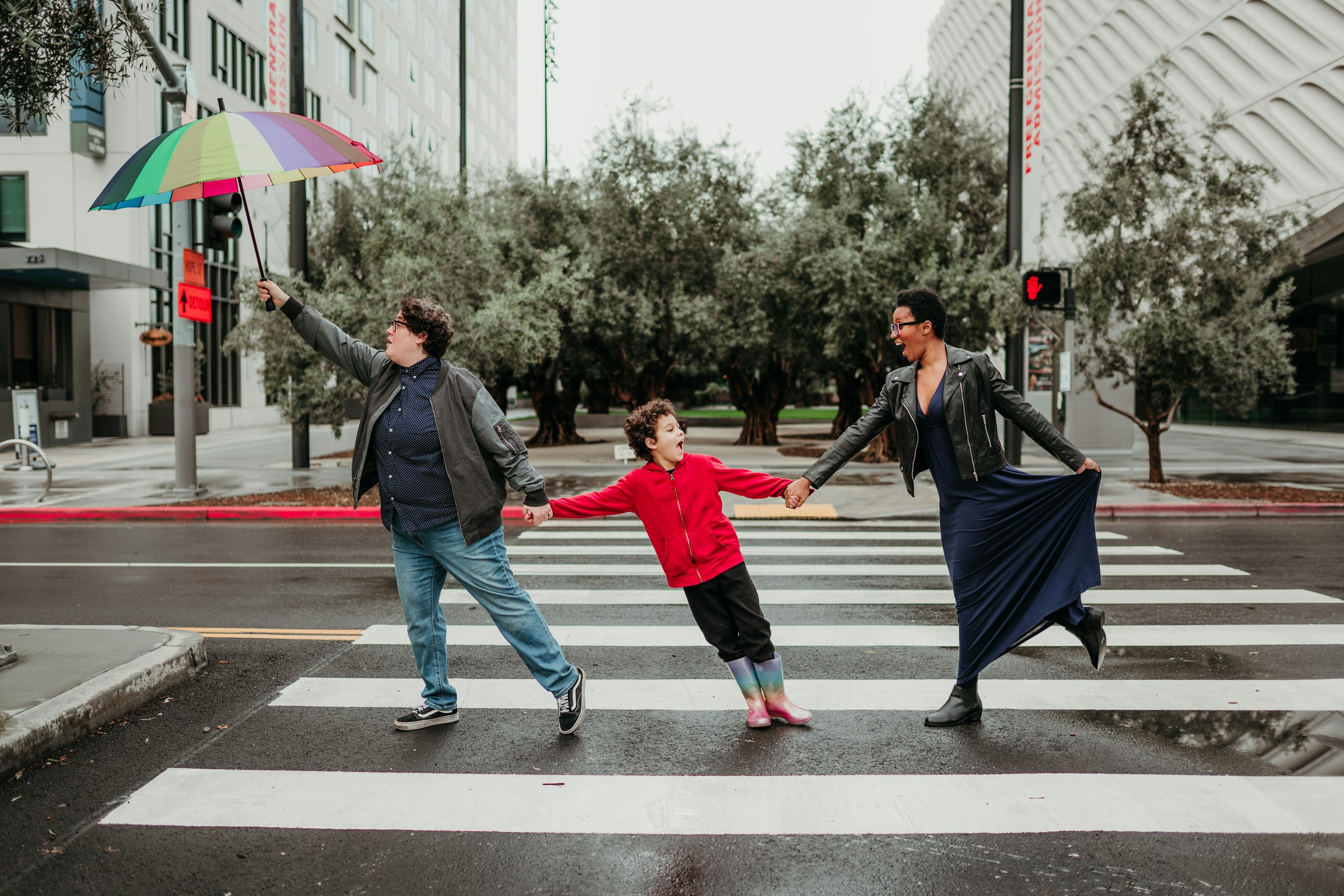Three people holding hands and crossing a city street while one person holds a colorful umbrella aloft.