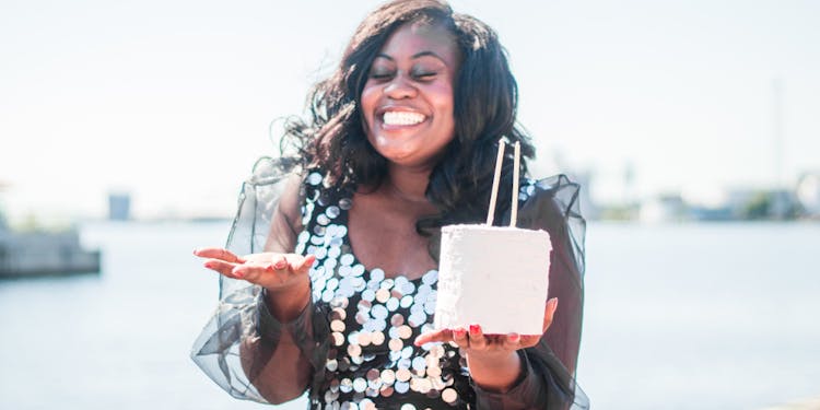 Happy woman in a sequined dress holding a small tiered cake outdoors near water.