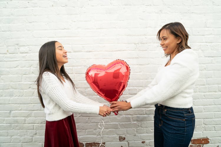 Two smiling people holding a red heart-shaped balloon against a white brick wall.