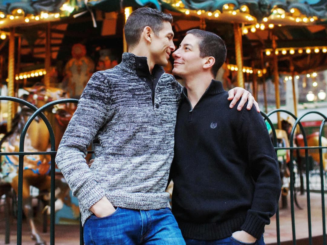 Two men affectionately leaning on each other in front of a carousel.