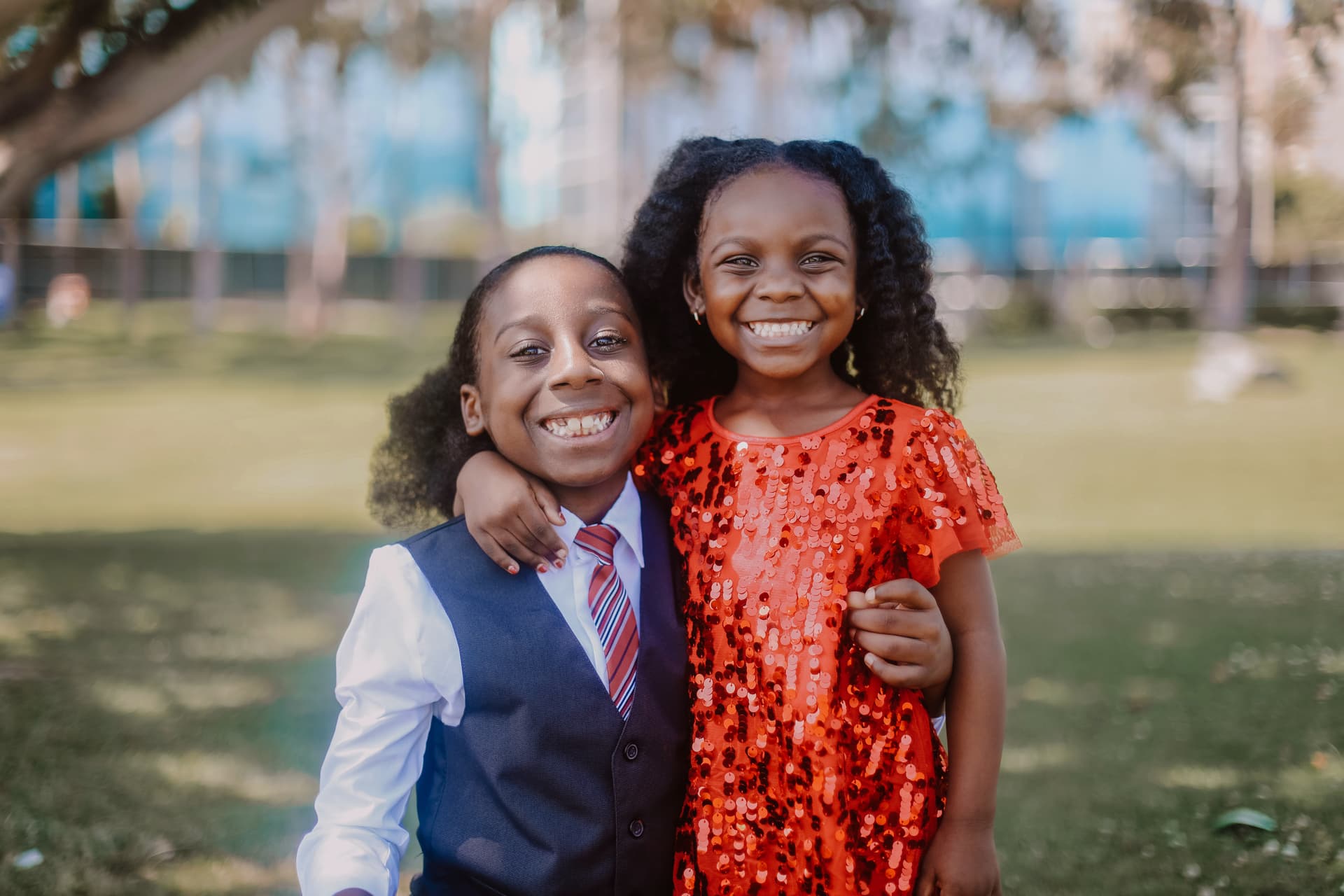 Two smiling children, a boy in a suit and a girl in a red dress, hugging each other in a park.