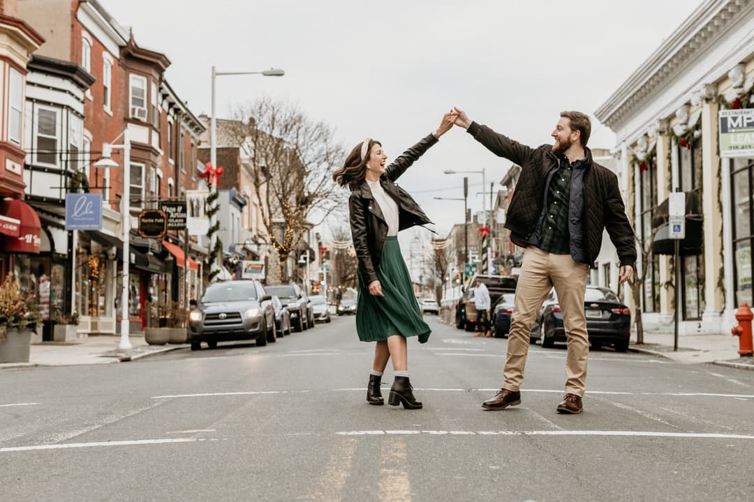 Couple dancing and holding hands in the middle of a city street during a Shoott outdoor photoshoot, with shops, cars, and city buildings in the background