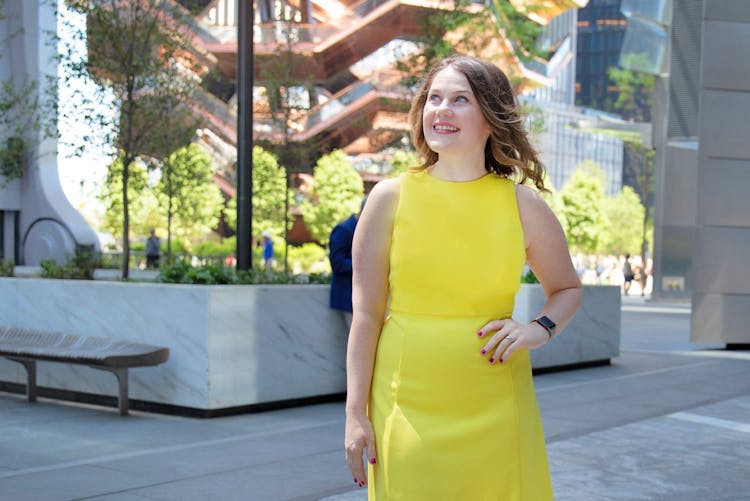 Woman in a yellow dress standing in an urban plaza, looking away from the camera, with greenery and modern architecture in the background