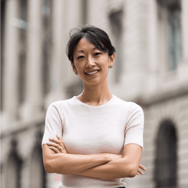A smiling woman with her arms crossed standing outdoors in a casual shirt with buildings in the background.