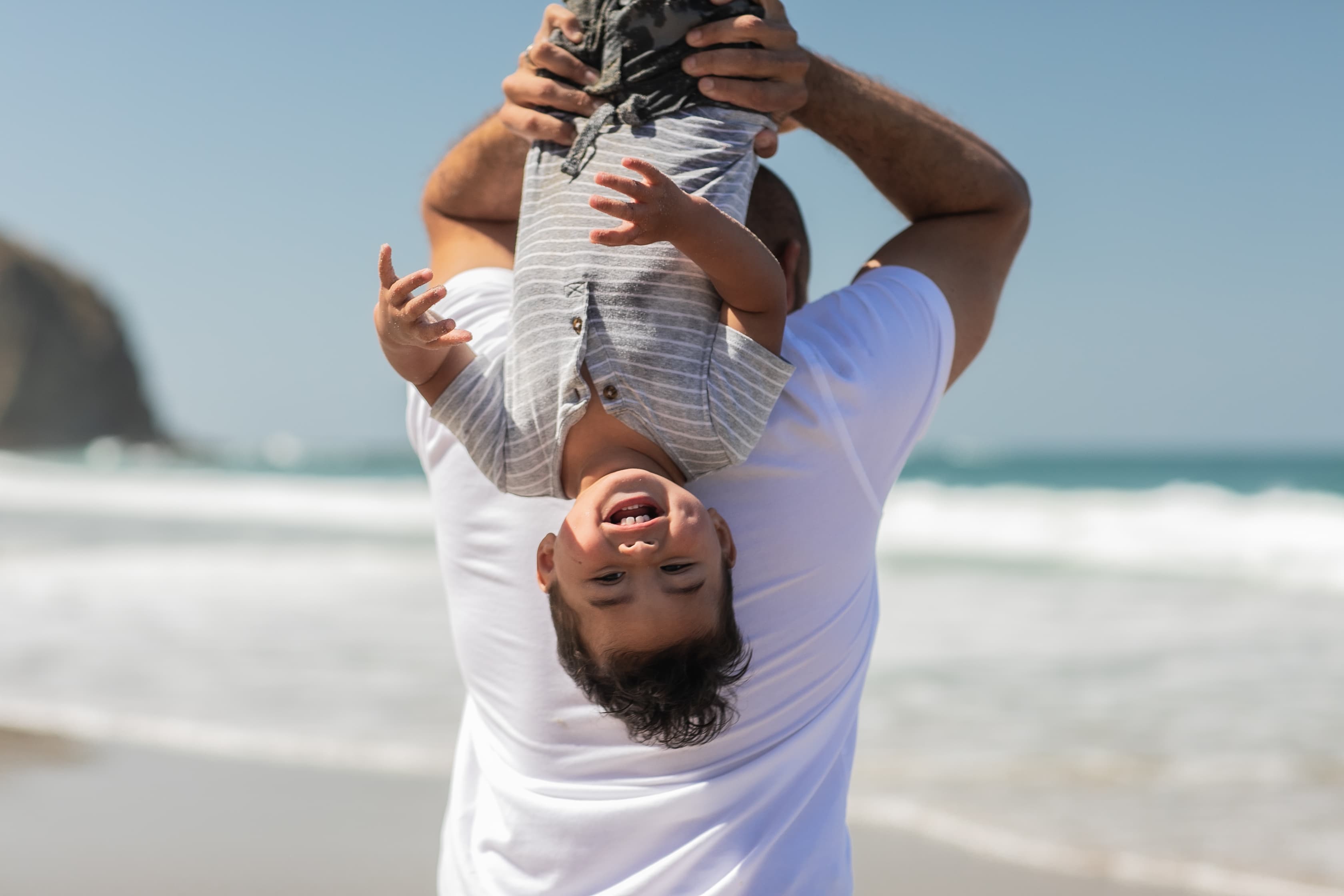 A man holding a laughing child upside down at the beach.