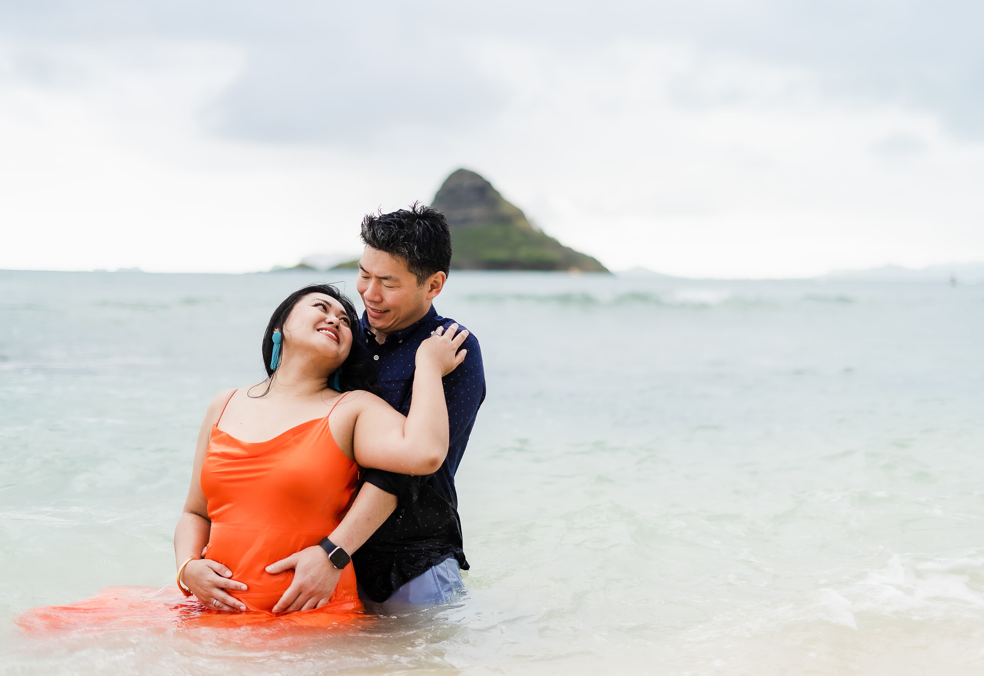 A couple embracing in shallow sea water with a mountain in the background.