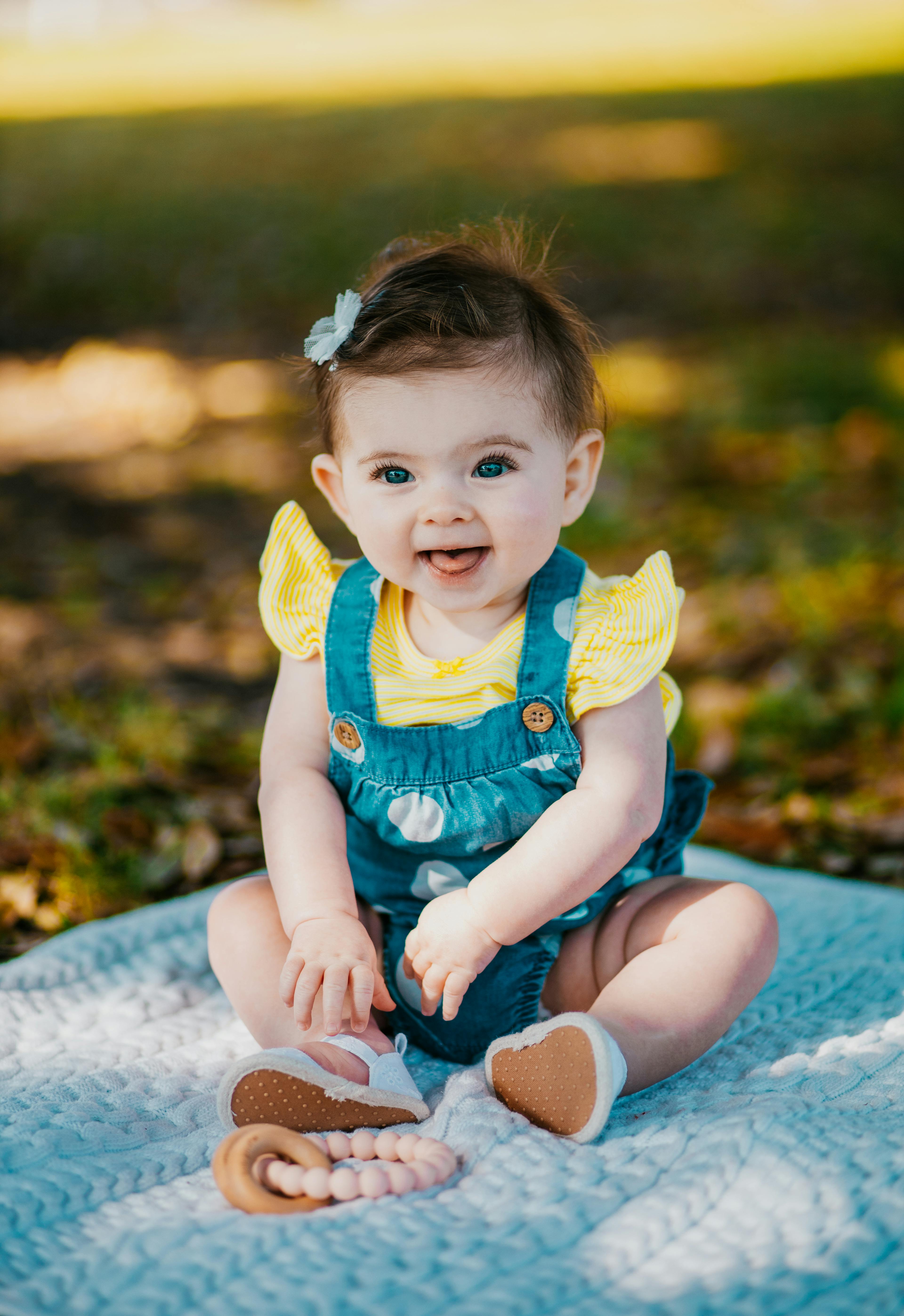 A cheerful baby sitting on a blue blanket on the grass with a bow in her hair