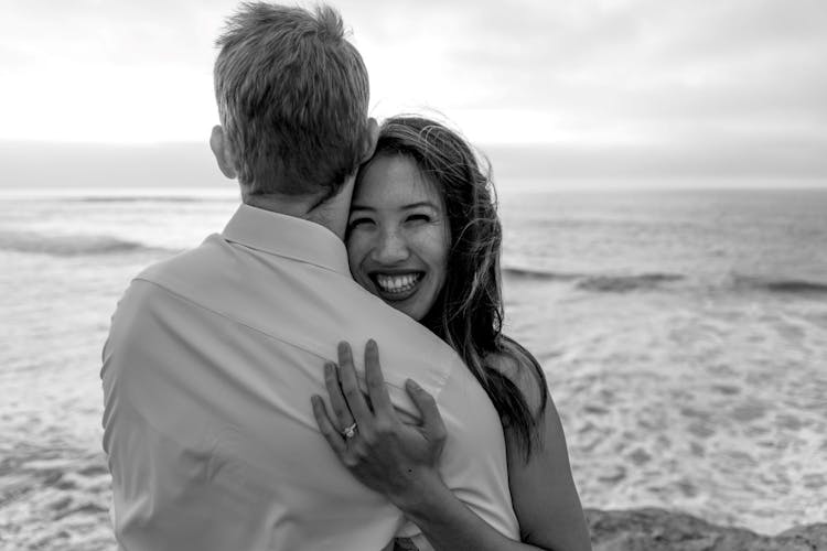 Woman hugging a man from behind at the beach, with a joyful smile