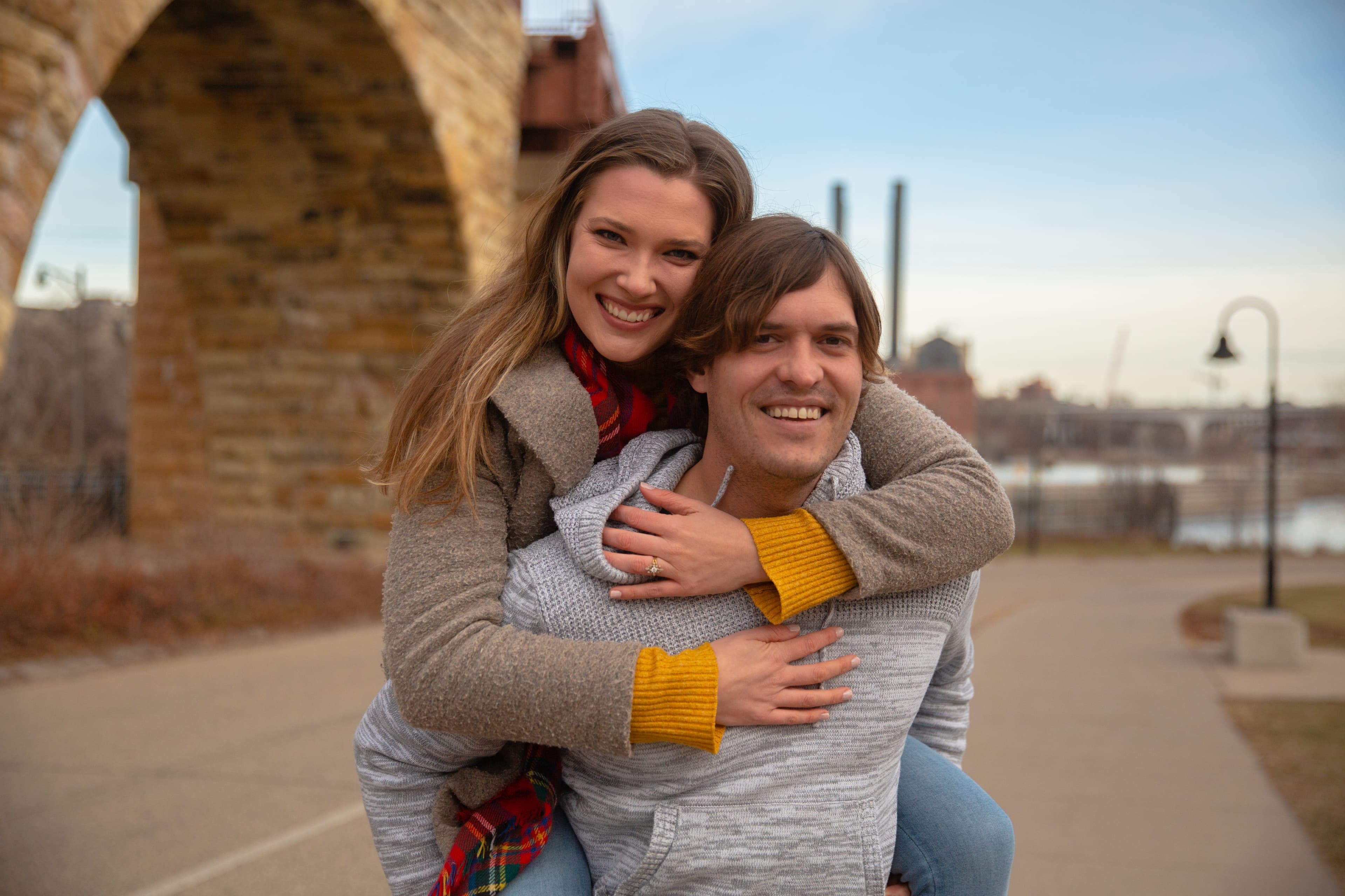 A smiling woman with her arms around a man's neck, piggybacking on him with an urban park backdrop.