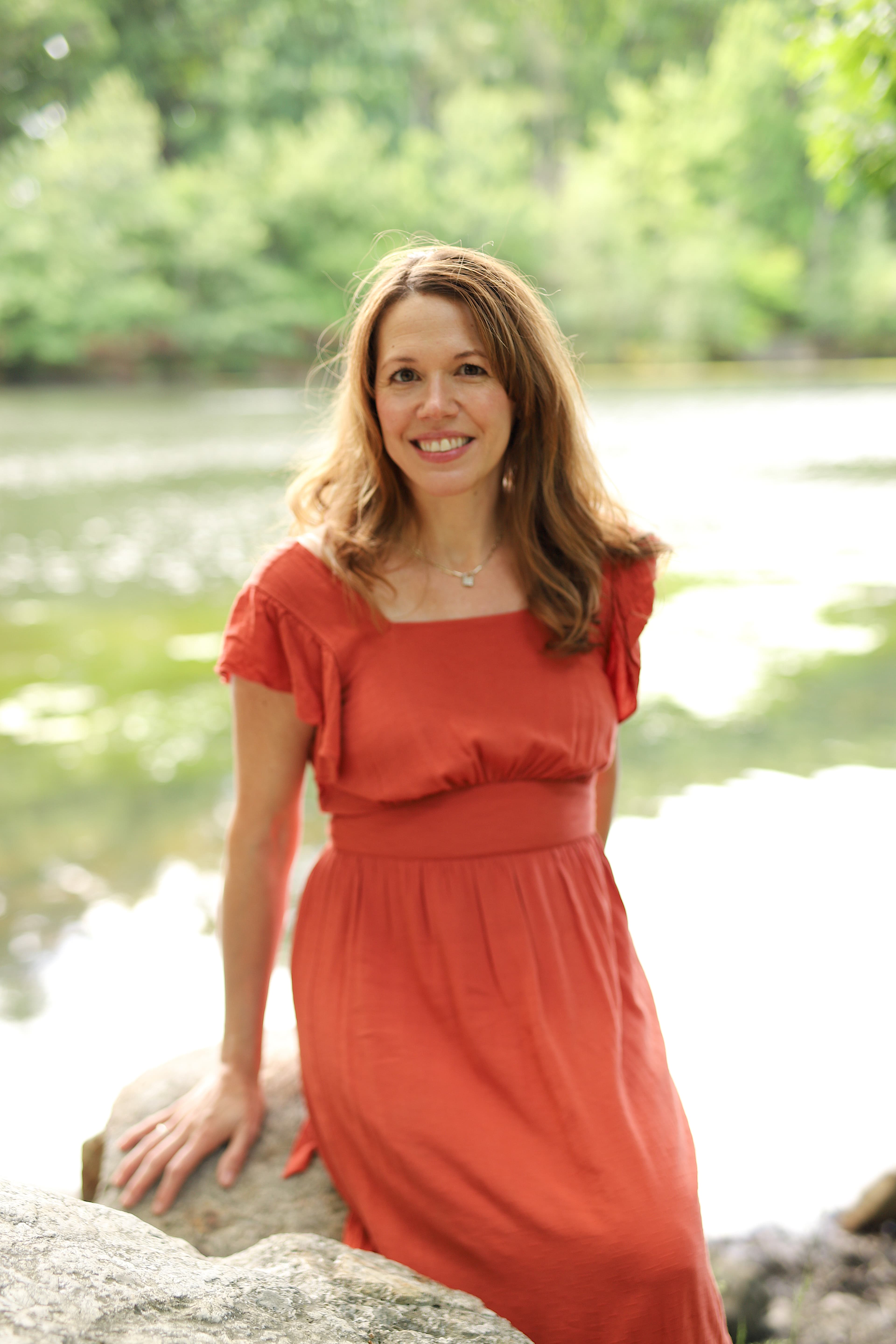 A woman in a red dress smiling while sitting on a rock by a pond.