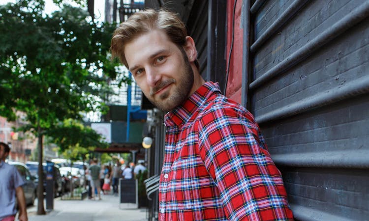 Man with a beard in a red plaid shirt leaning against a dark wall