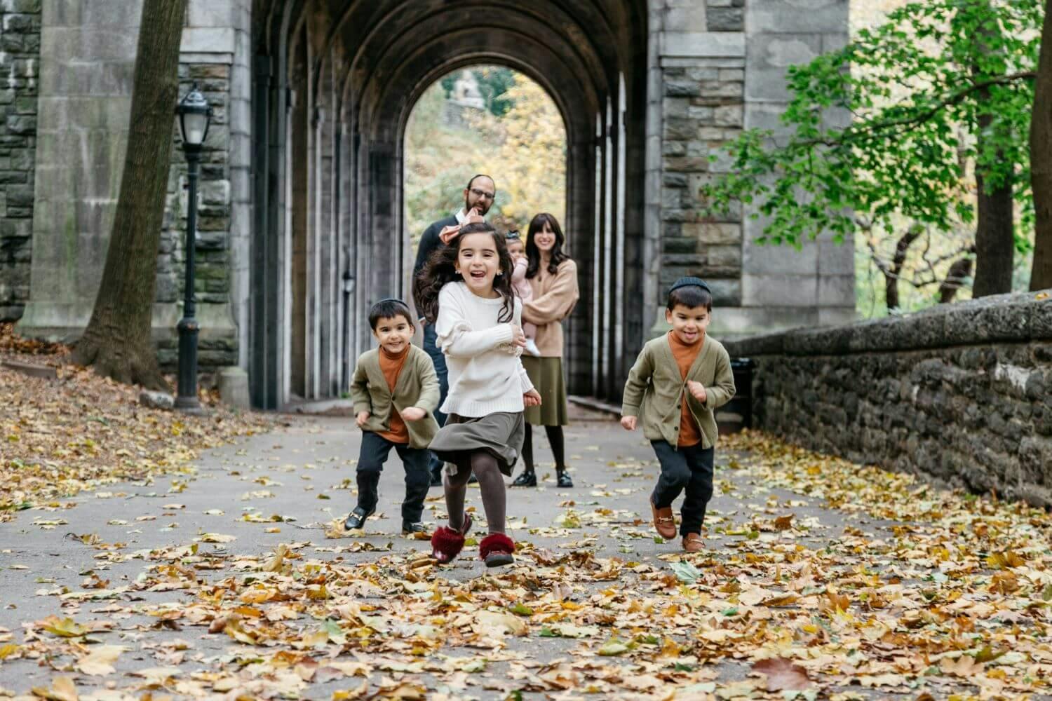Two children happily running towards the camera with three adults following behind, in a scenic autumn archway.
