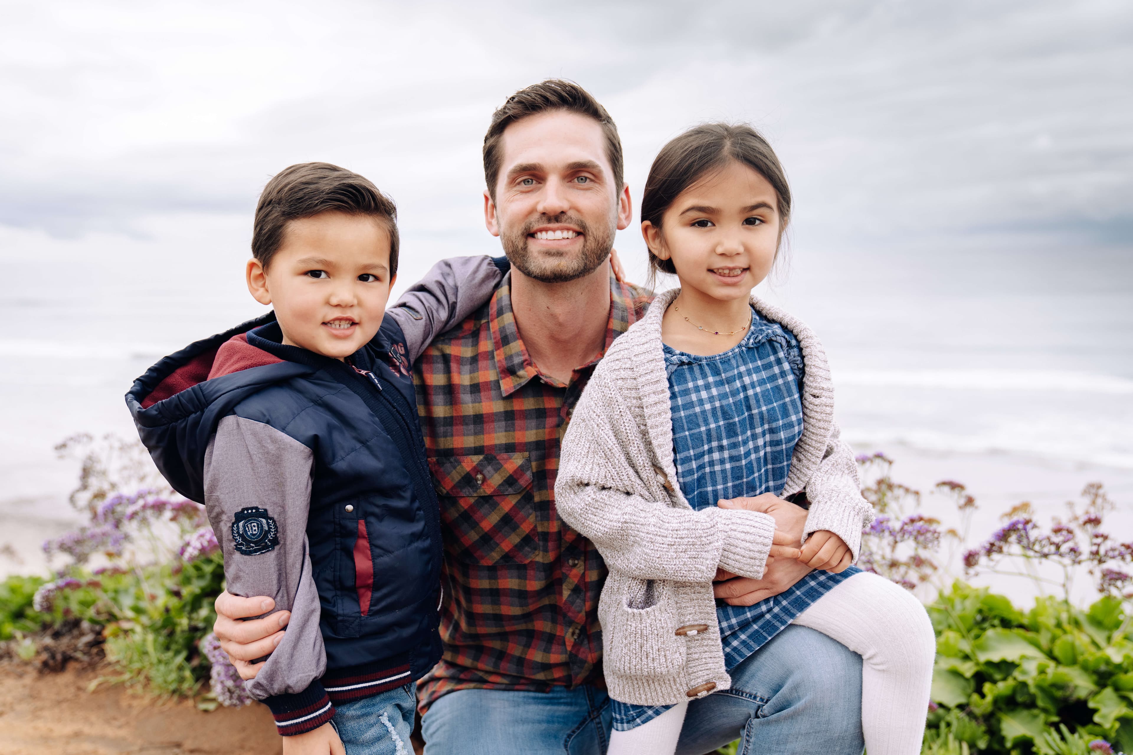 A man with two young children smiling at the camera with a cloudy beach in the background