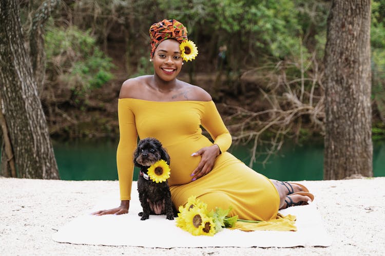 Pregnant woman in a yellow dress sitting with a small black dog surrounded by sunflowers