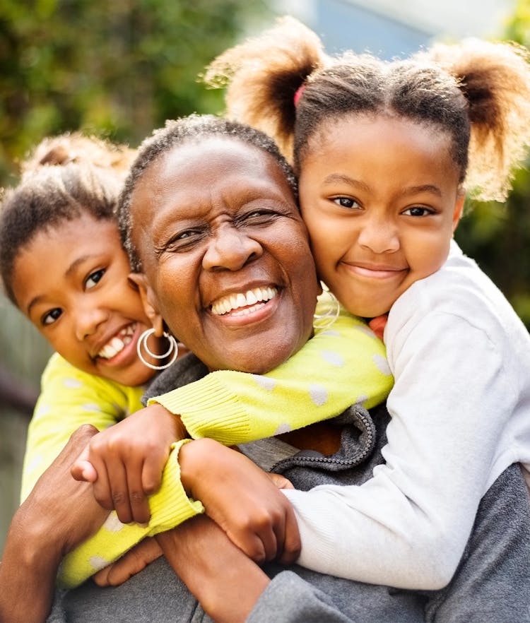A joyful grandmother hugging her two smiling grandchildren outdoors.