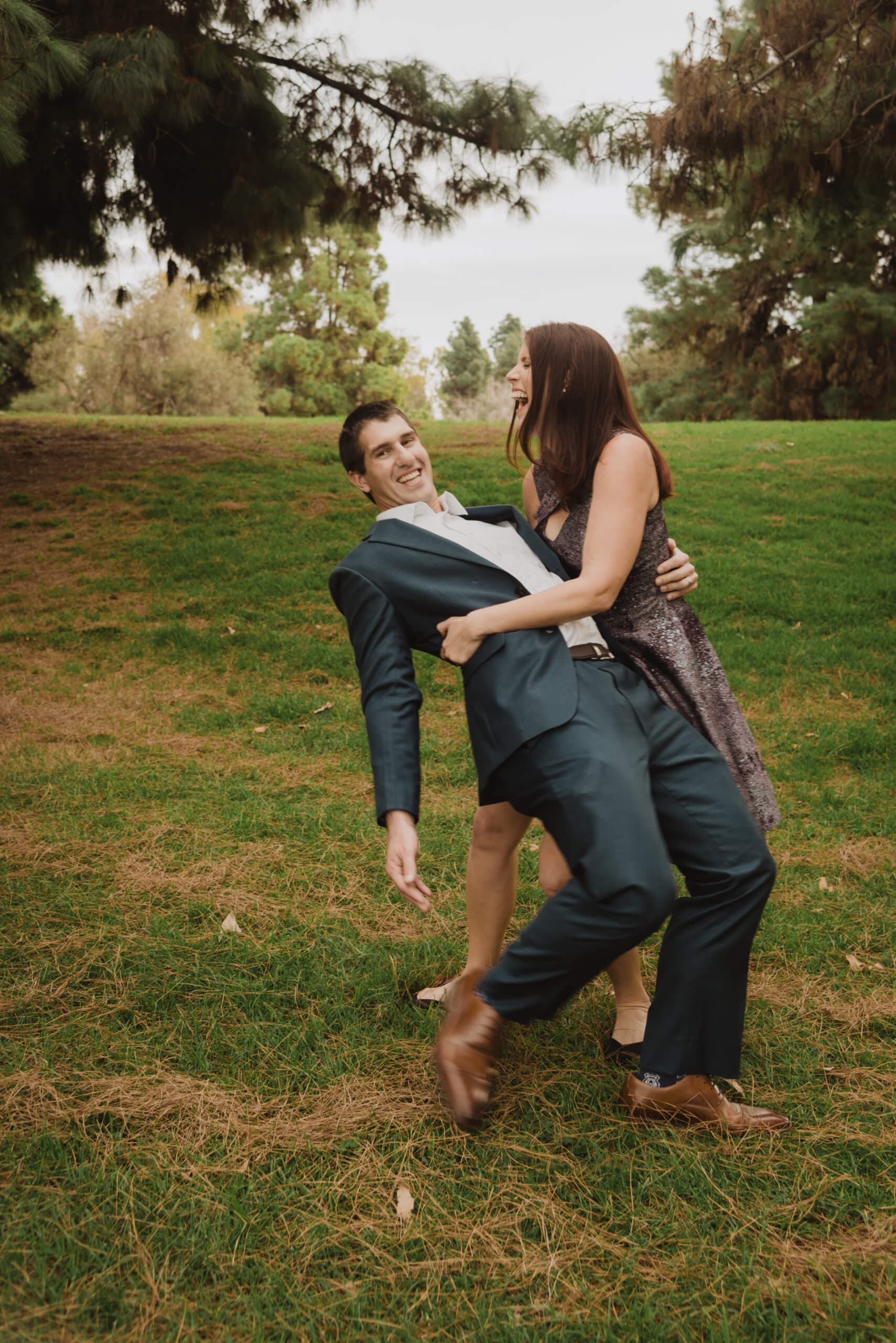 A couple playfully dancing on a grass field, with the man lifting the woman slightly off the ground