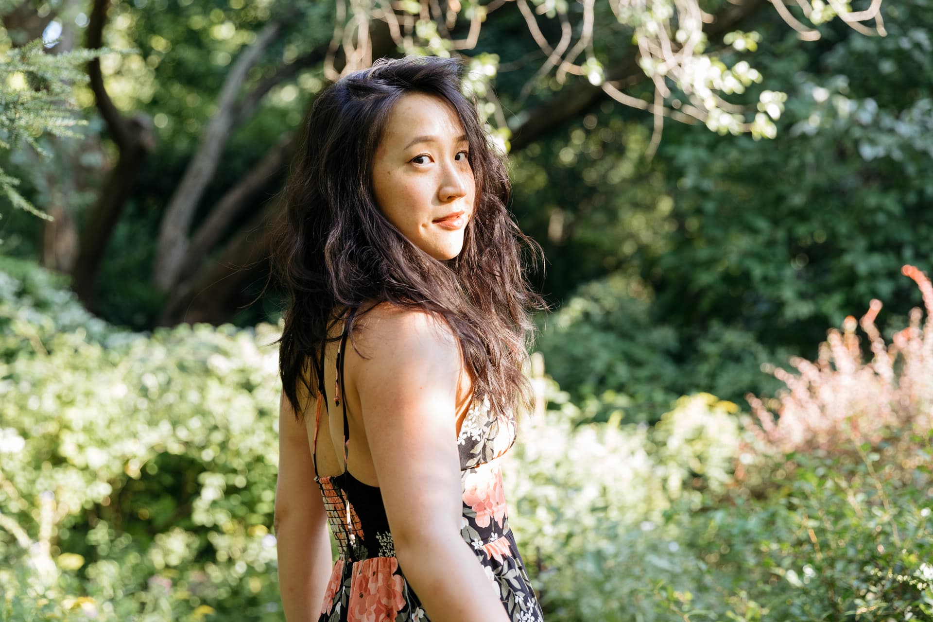A woman in a floral dress looking over her shoulder in a sunlit garden.