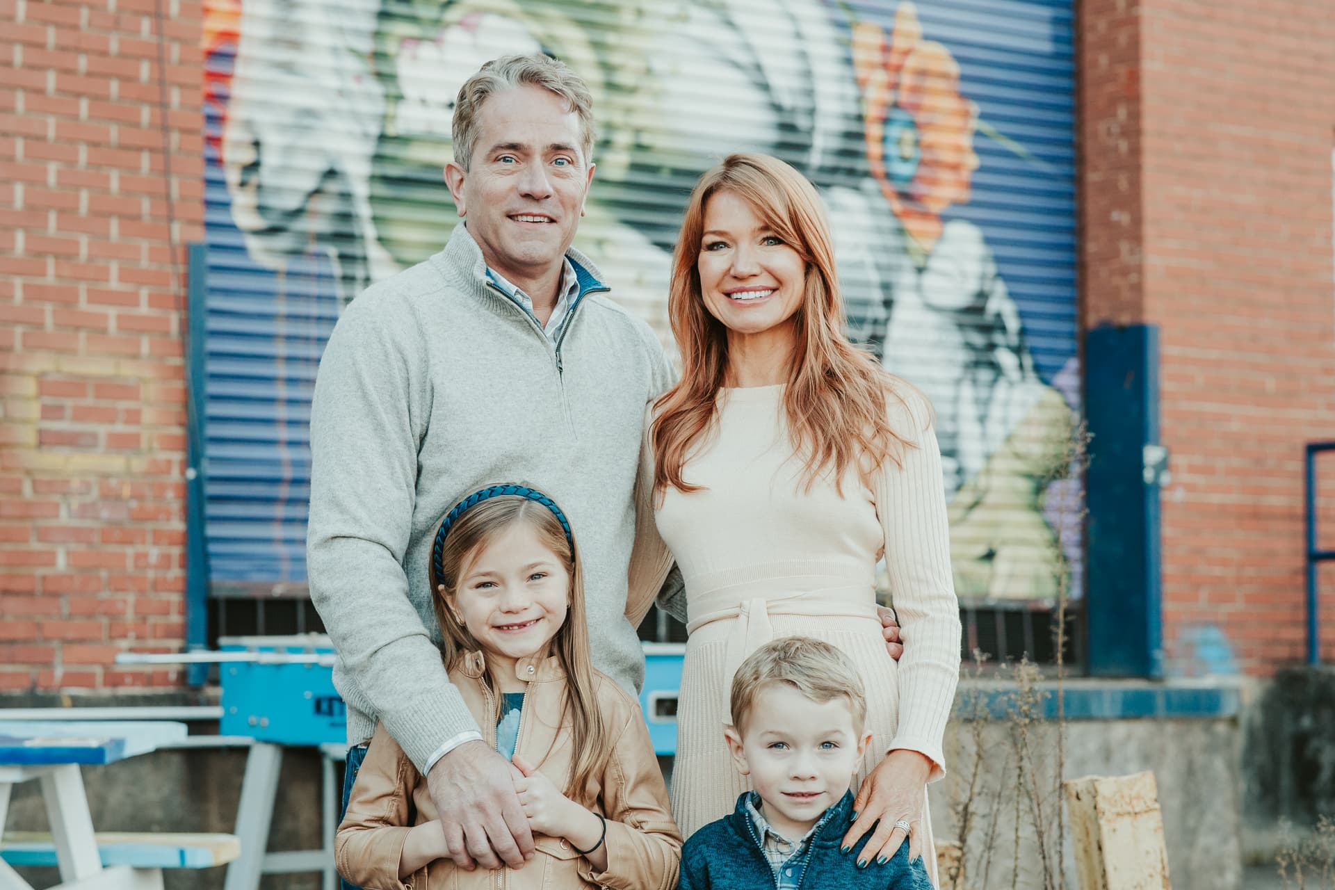 Family of four smiling outdoors in front of a colorful mural during a Shoott mini photoshoot, with mom, dad, daughter, and son posed together against a brick and mural backdrop.