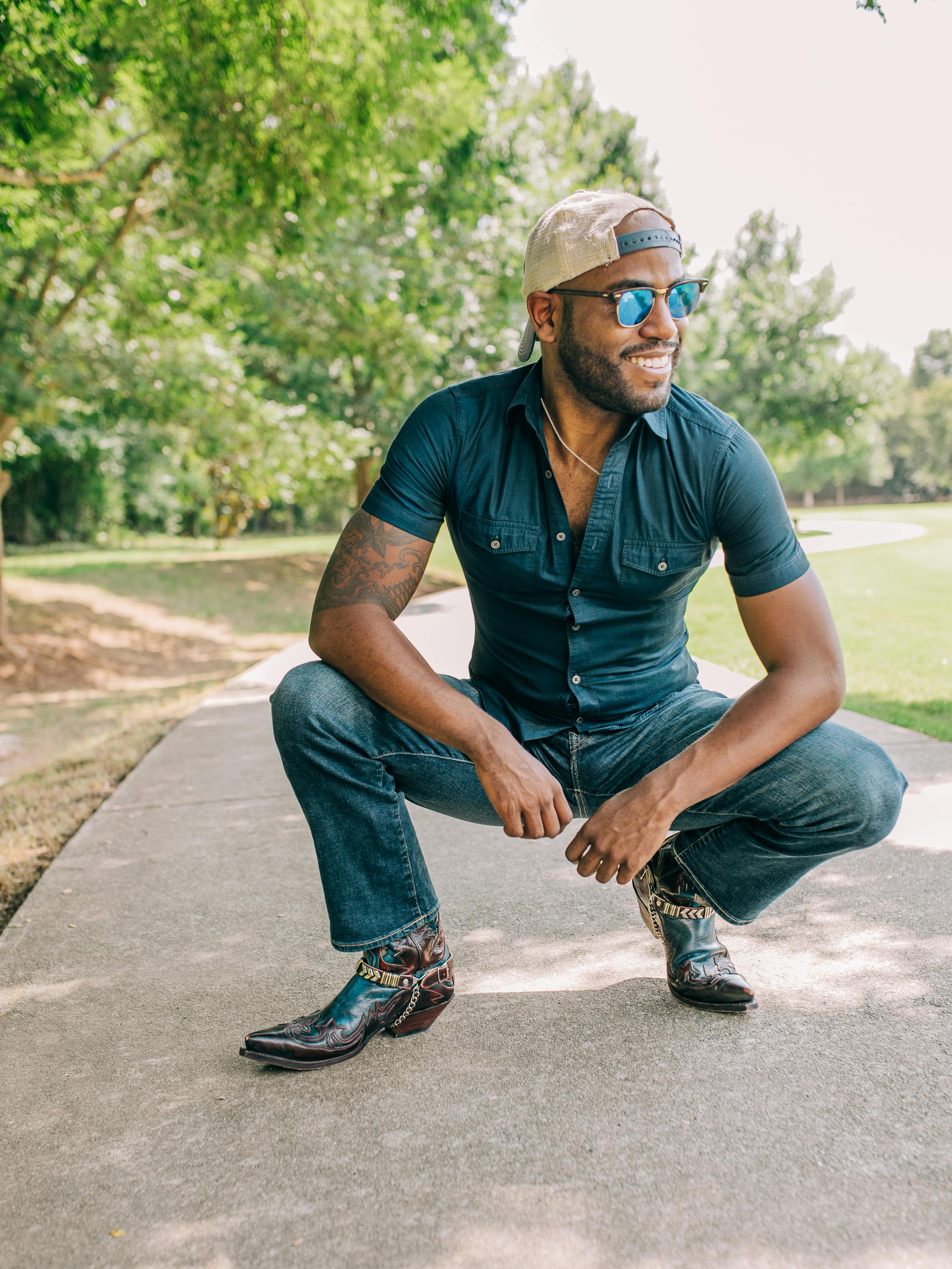 Man squatting on a sidewalk in a park, wearing sunglasses, a baseball cap, and cowboy boots.