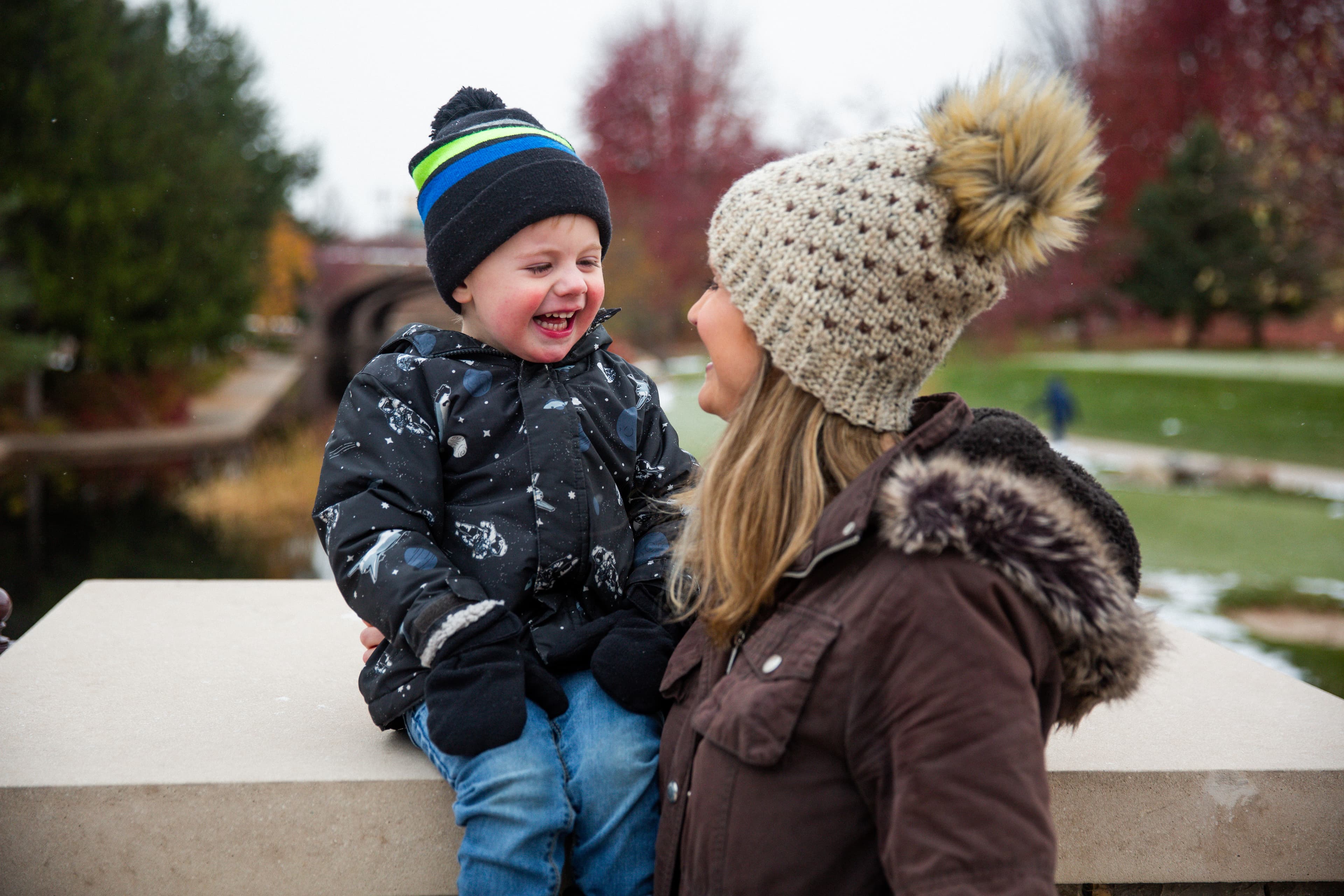 A joyful toddler in a winter hat laughing with an adult wearing a beanie in a park.