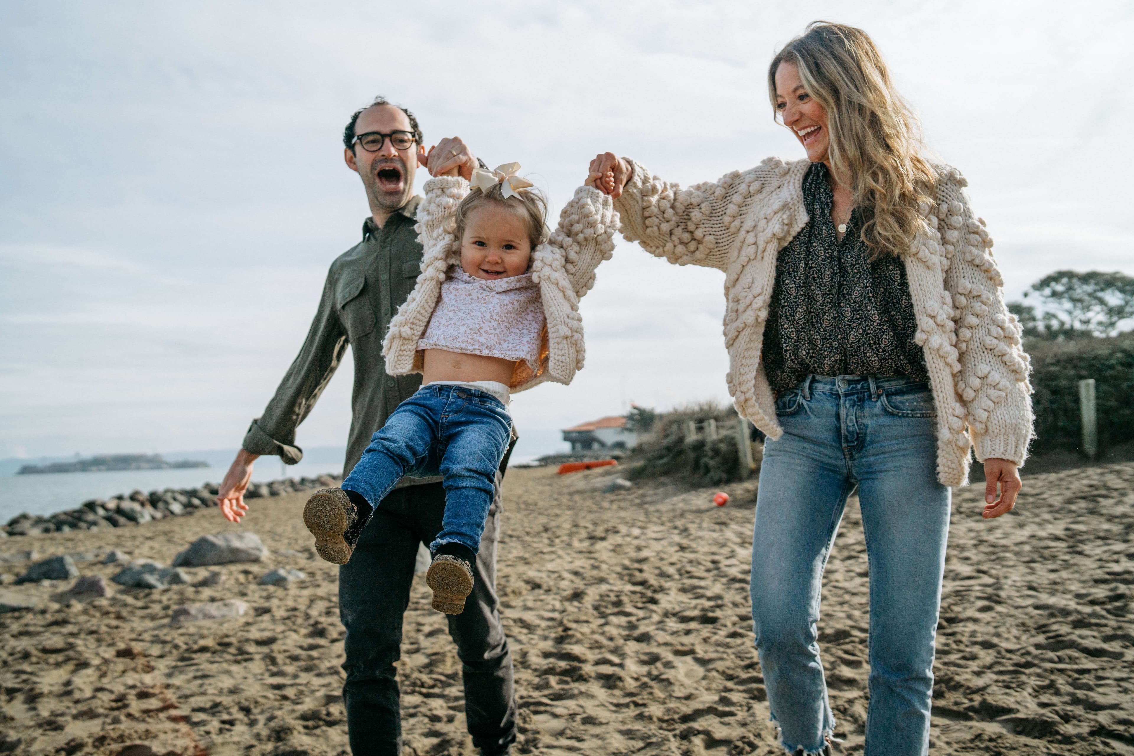 A family swinging a toddler by the arms on the beach with the ocean in the background.