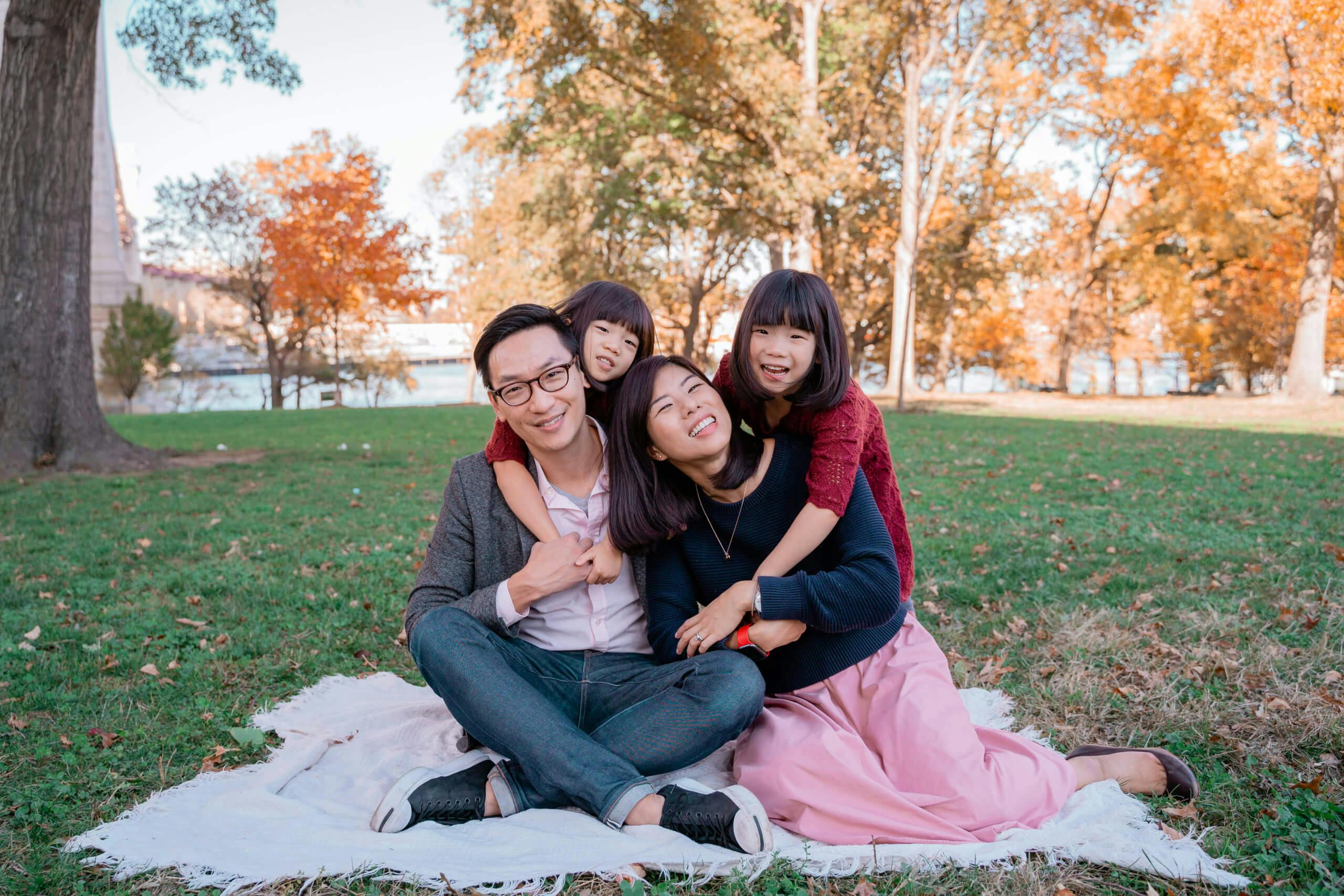 A family of four sitting on a blanket in a park with autumn leaves