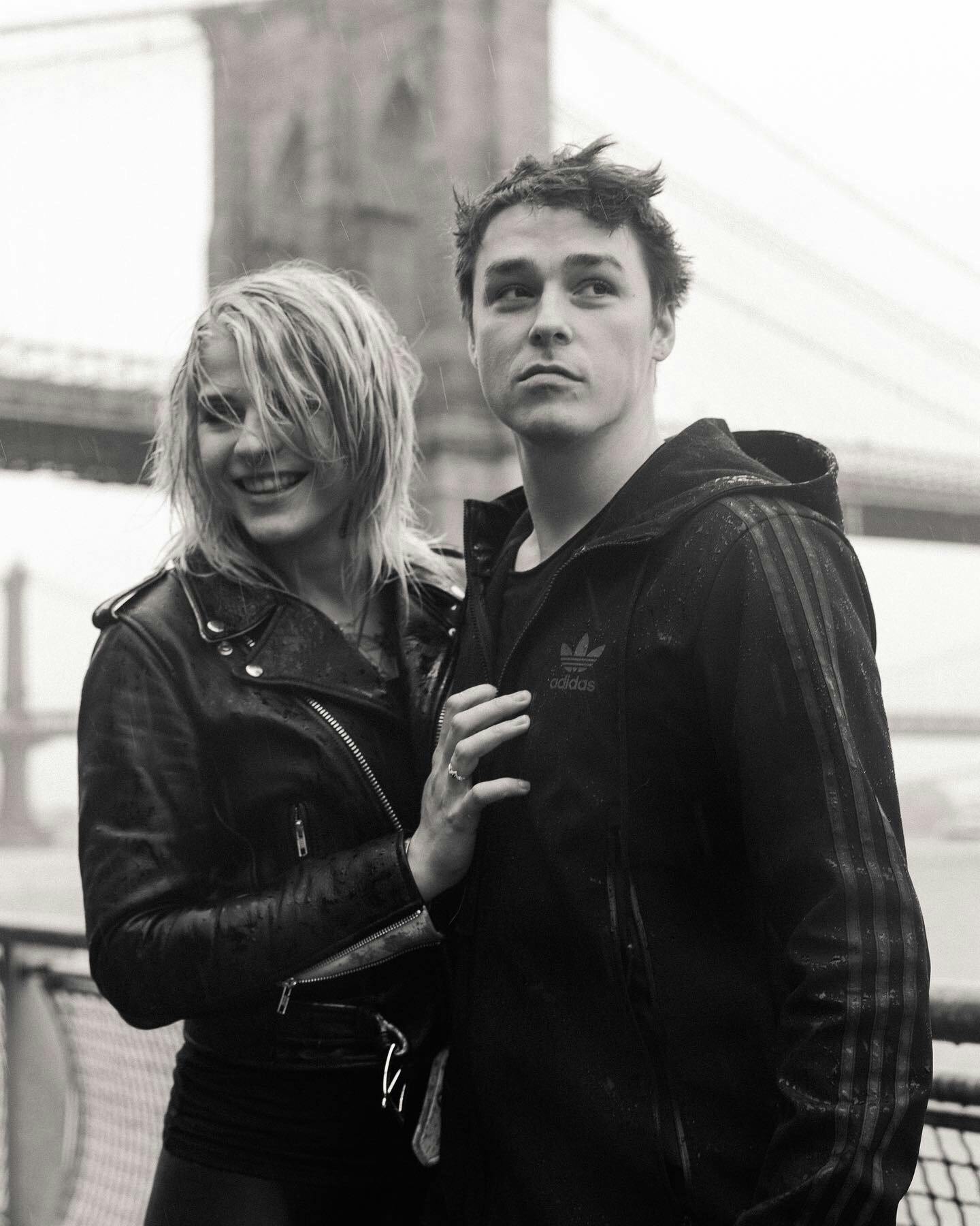 A young woman and man standing in front of a bridge, both with wet hair and leather jackets.