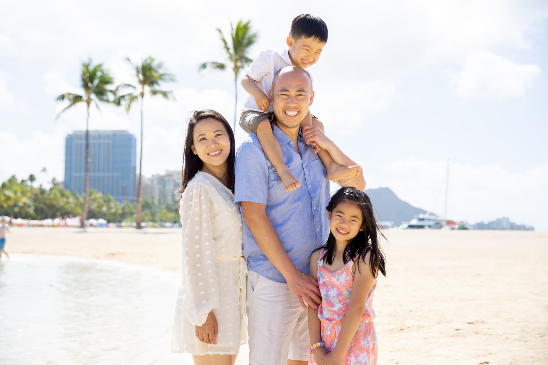 Family of four smiling on a sunny Waikiki beach, with the father carrying his young son on his shoulders and Diamond Head visible in the background, captured during a Shoott family photoshoot.
