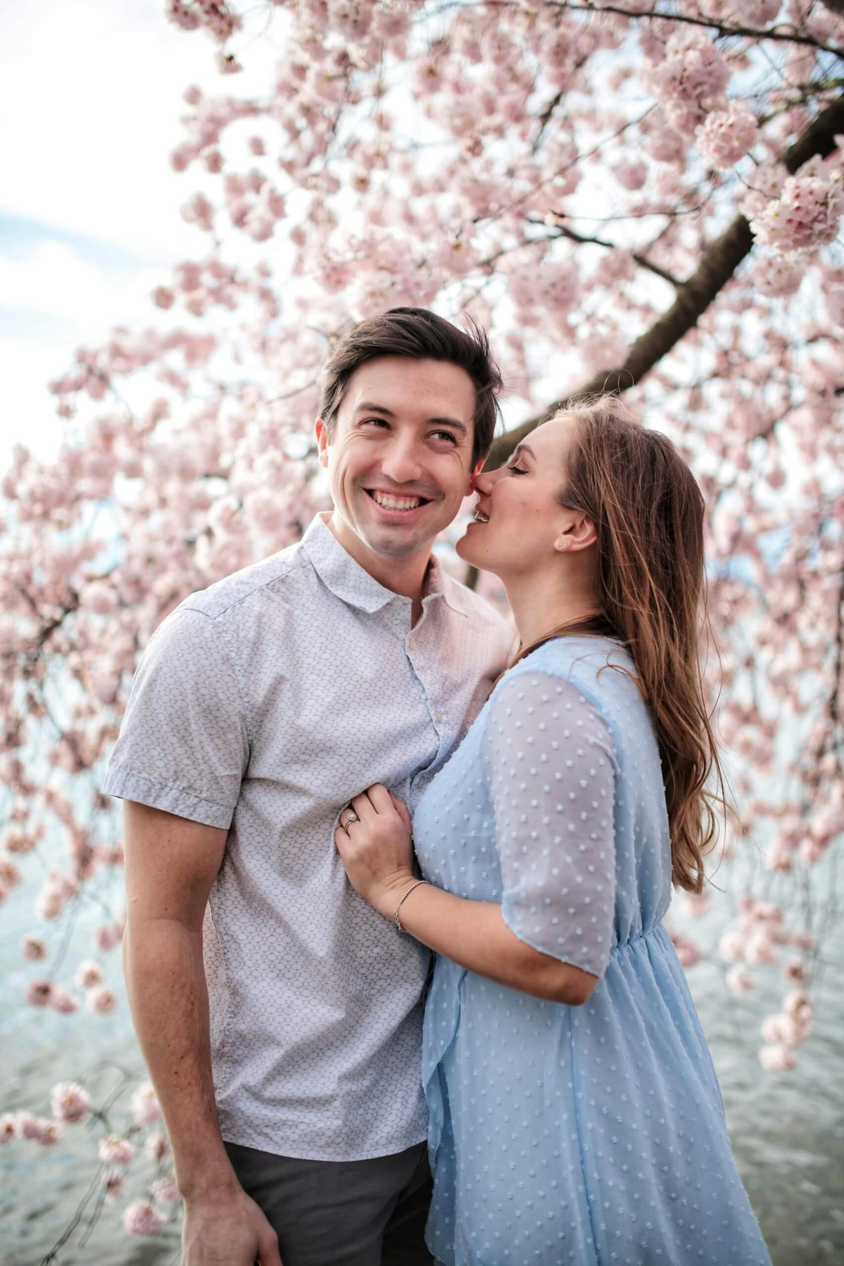 A couple with a woman kissing a man on the cheek under cherry blossom trees.