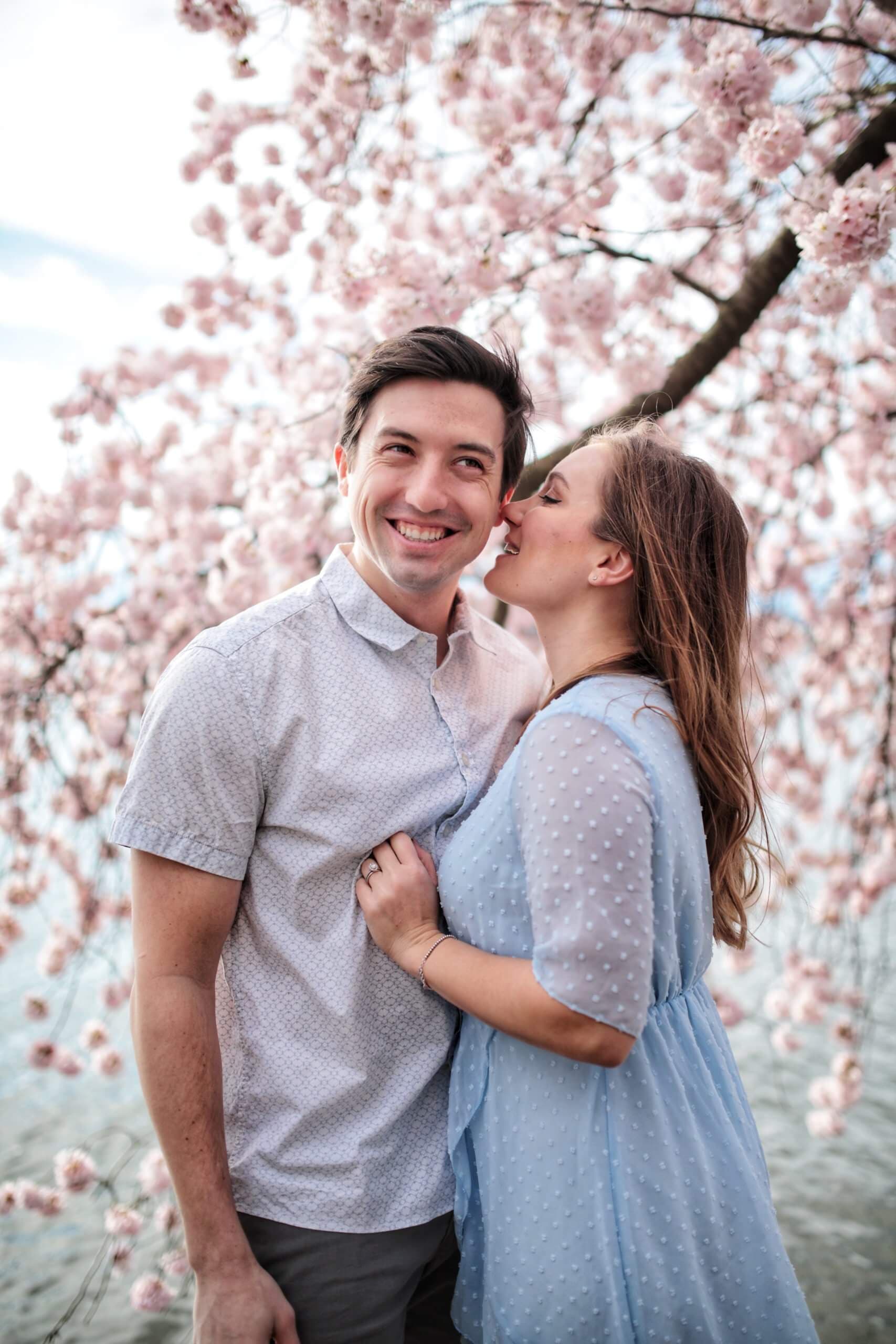 A couple with a woman kissing a man on the cheek under cherry blossom trees.