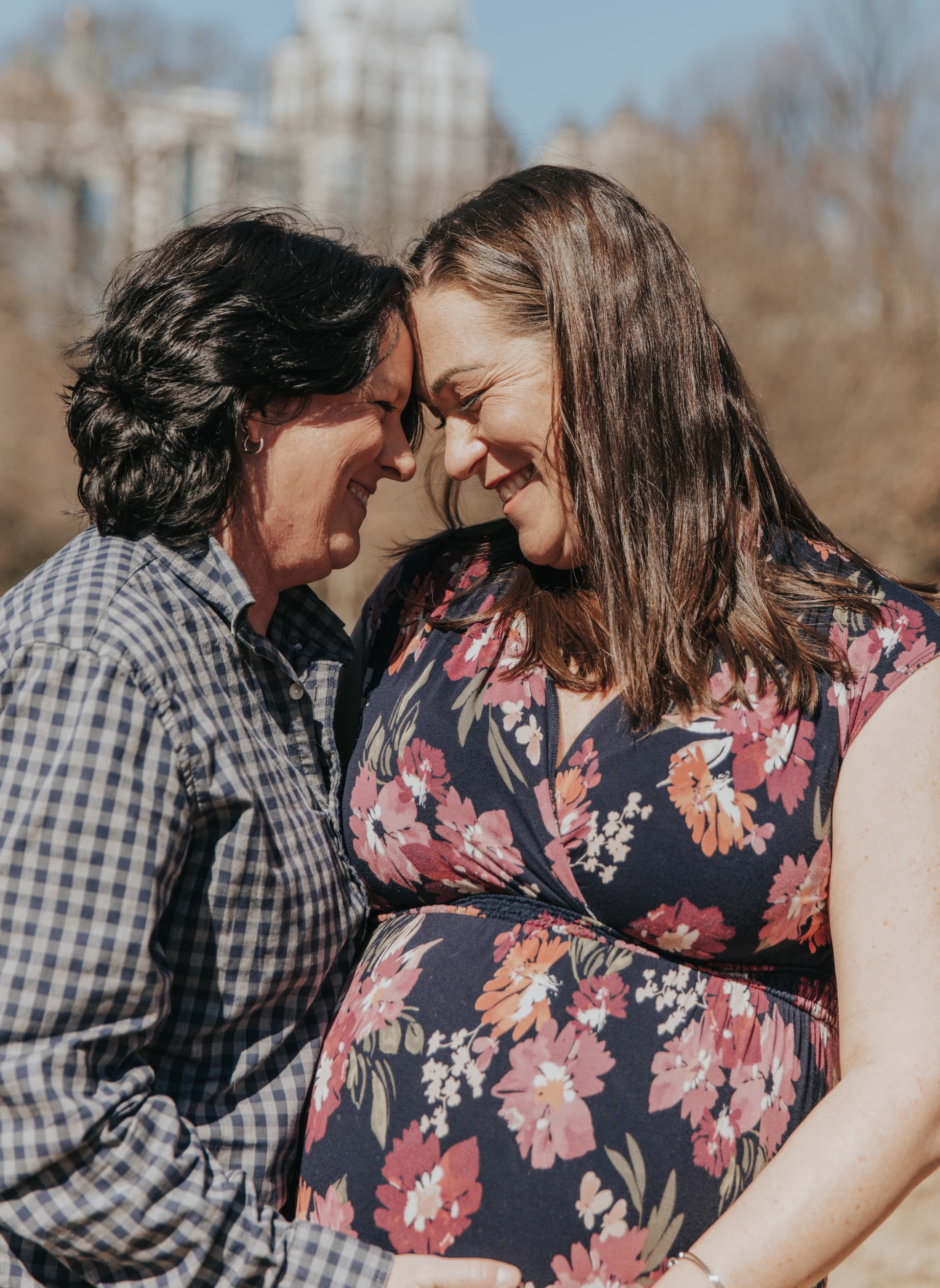 Two smiling women with foreheads touching, one pregnant, in a sunny outdoor setting.