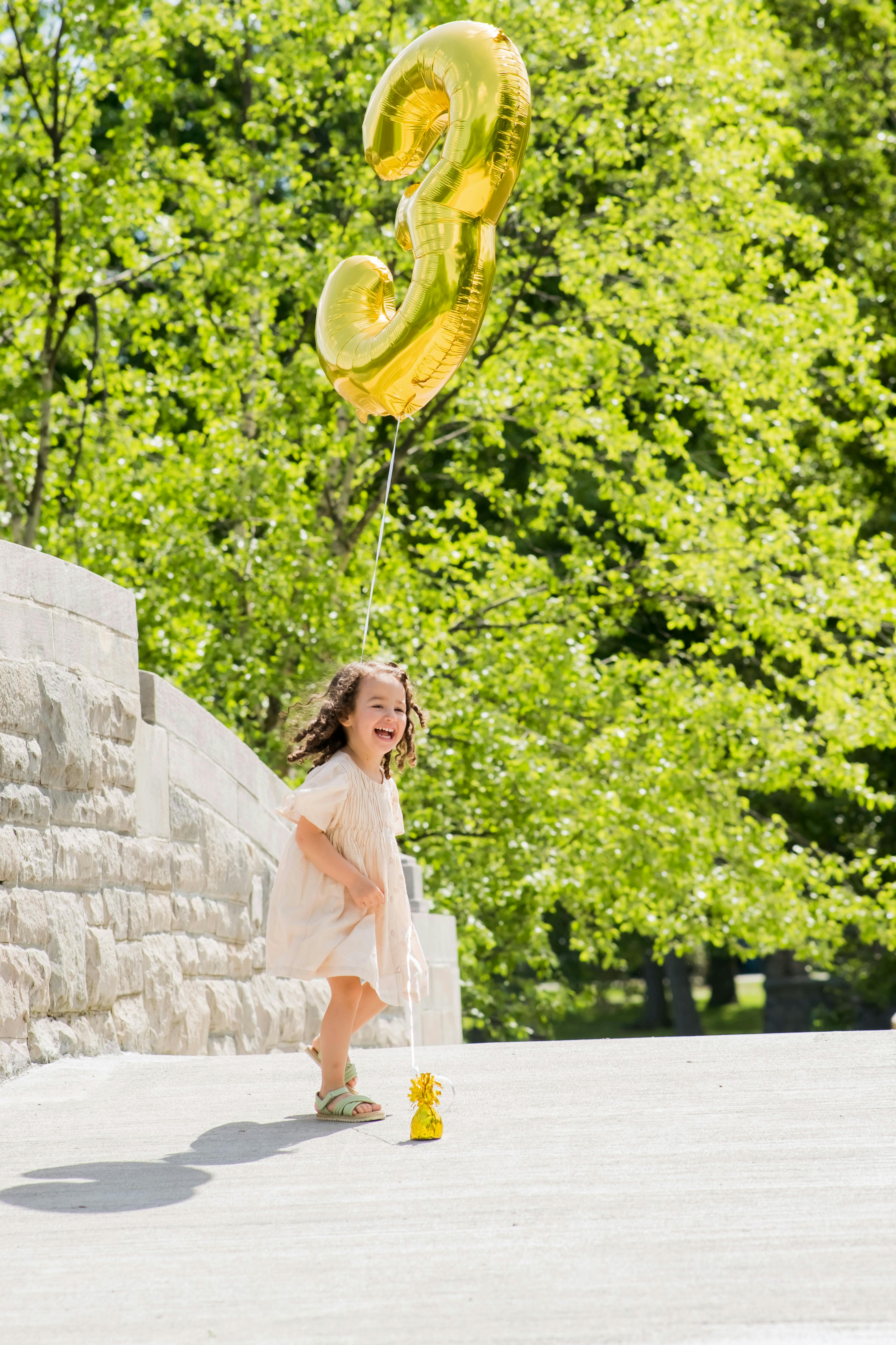 A young child happily holding a number 3 shaped balloon outdoors with a sunny green background.