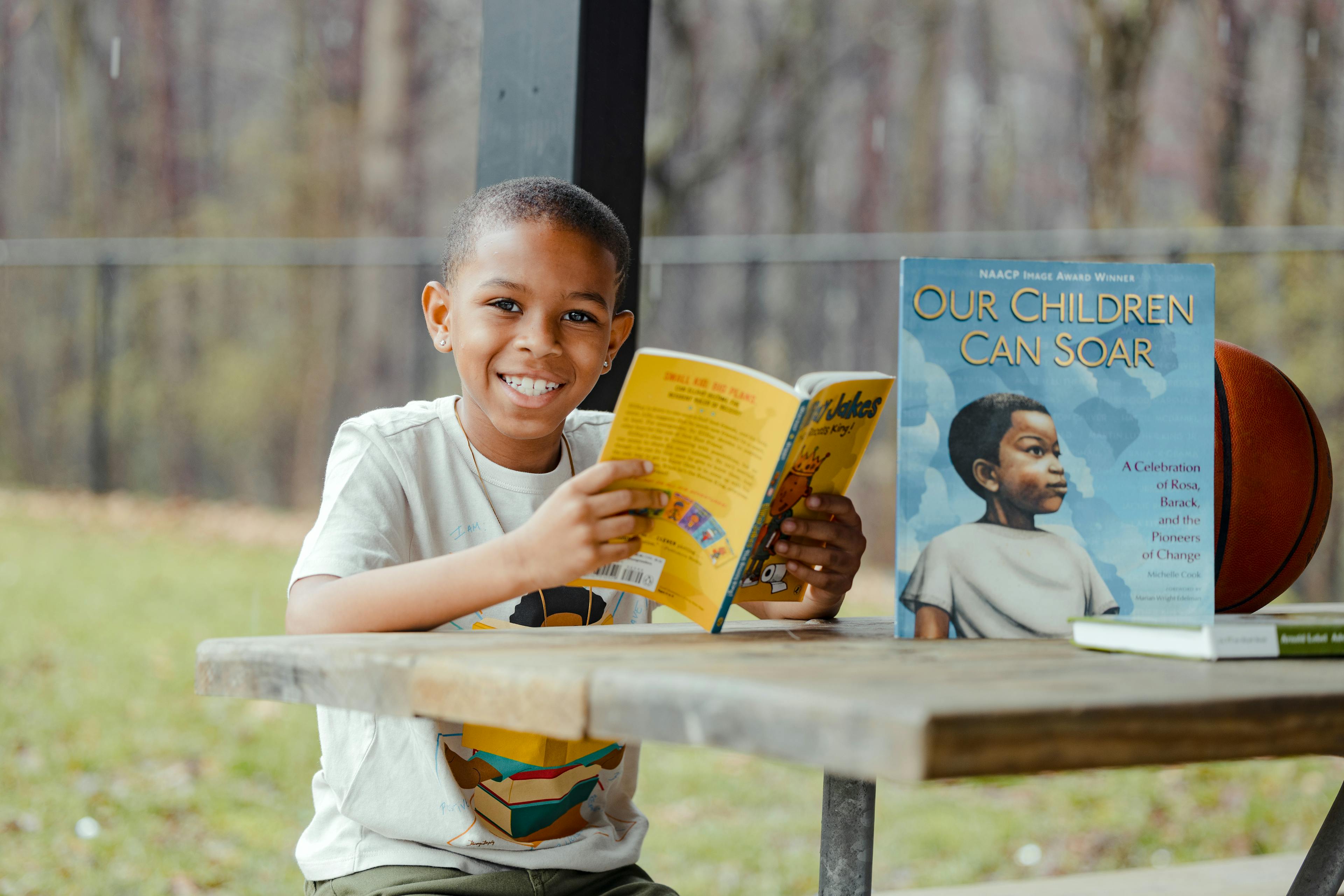 A smiling child reading a book at a picnic table with a basketball beside them.