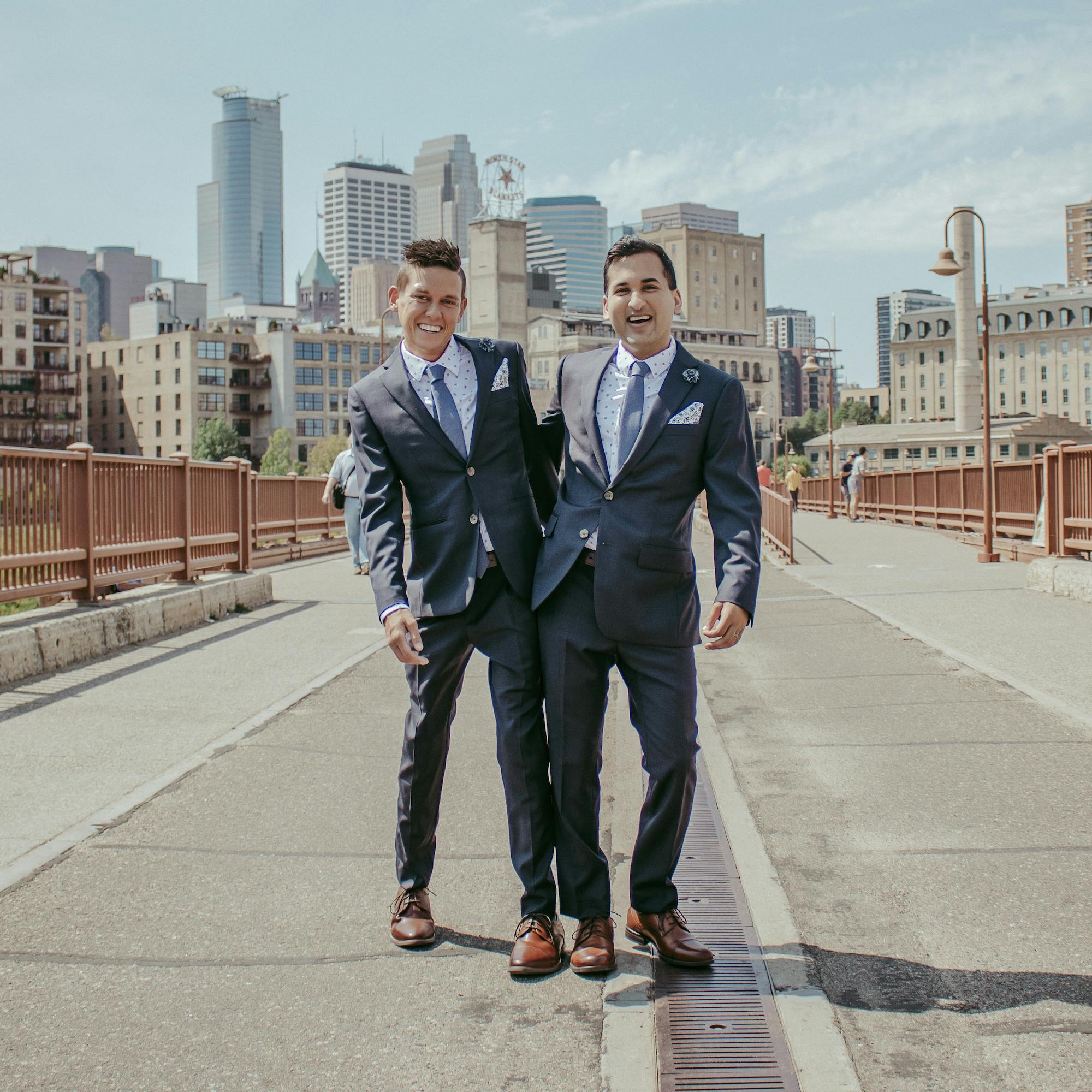Two smiling men in suits standing on a bridge with a city skyline in the background.
