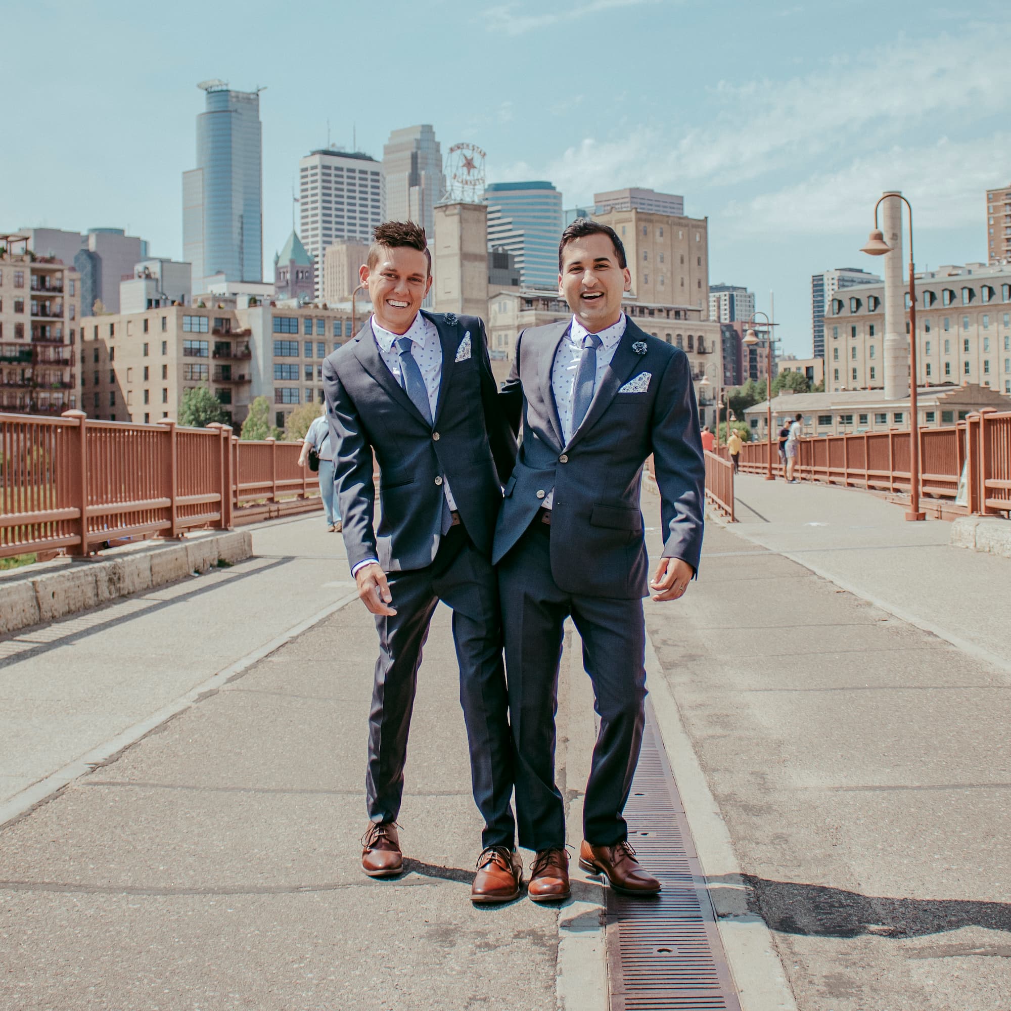 Two smiling men in suits standing on a bridge with a city skyline in the background.