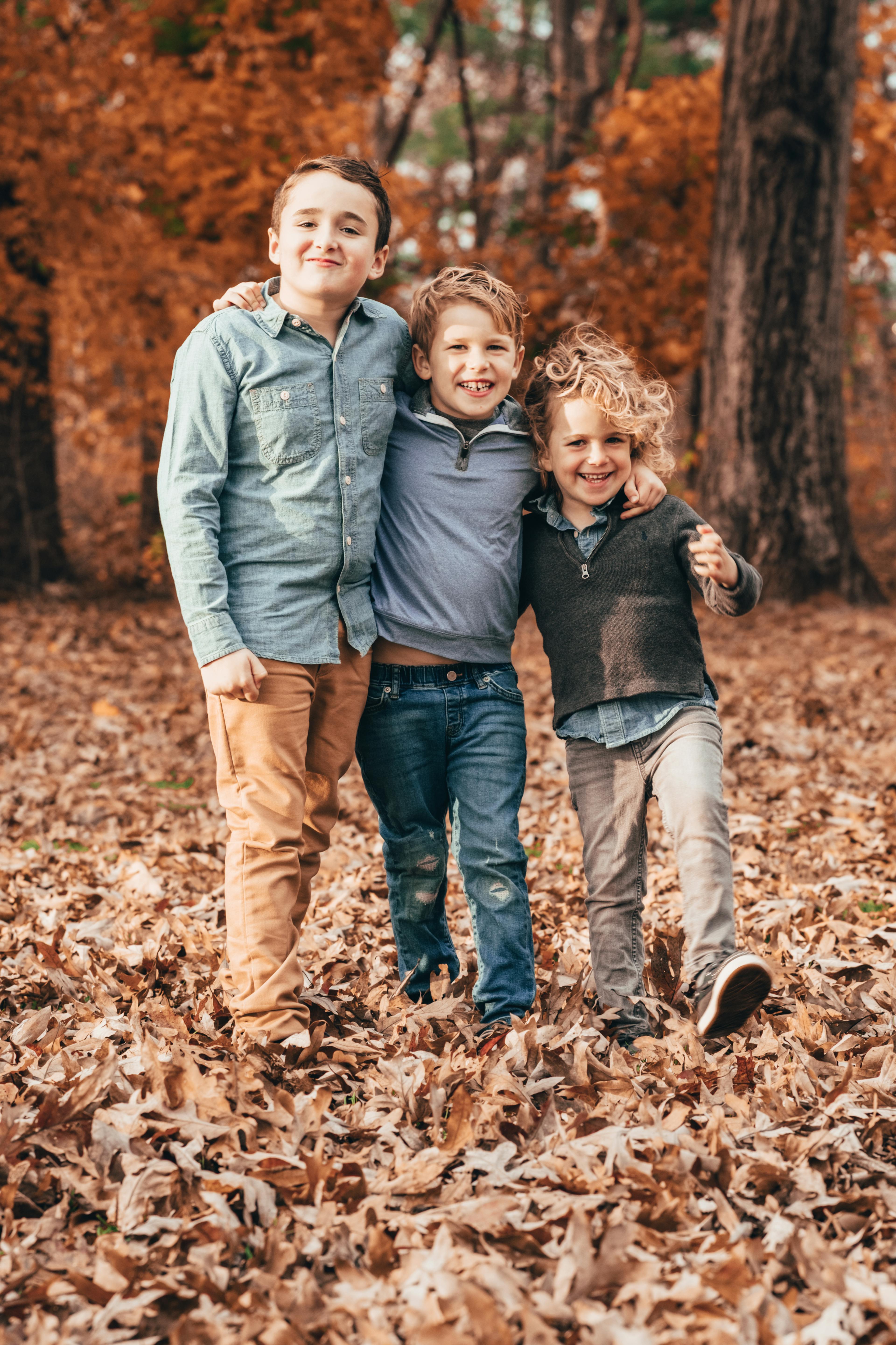 Three boys hugging and smiling among fallen leaves in an autumn setting.
