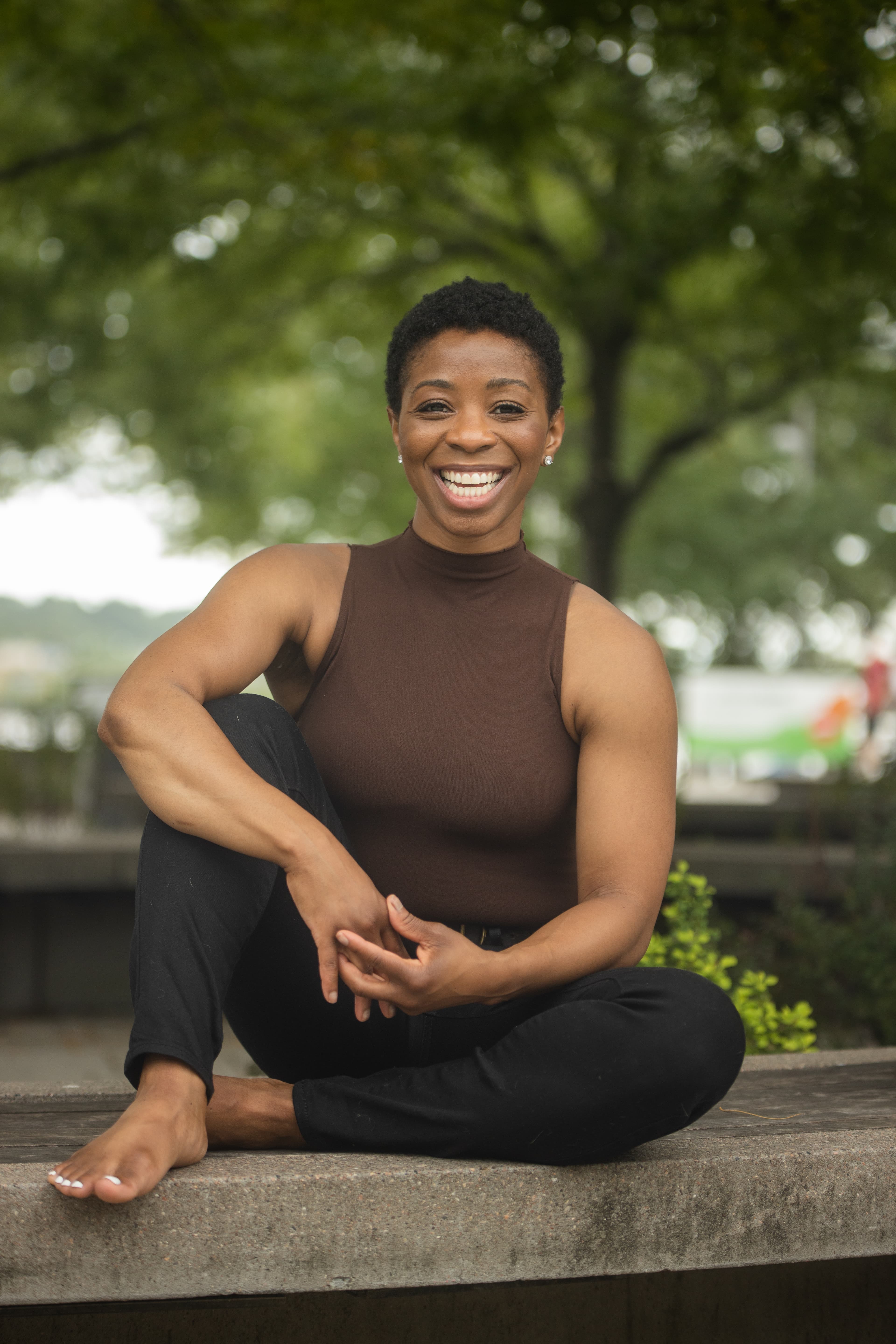 Smiling woman in a sleeveless top sitting cross-legged on a stone ledge in a park.