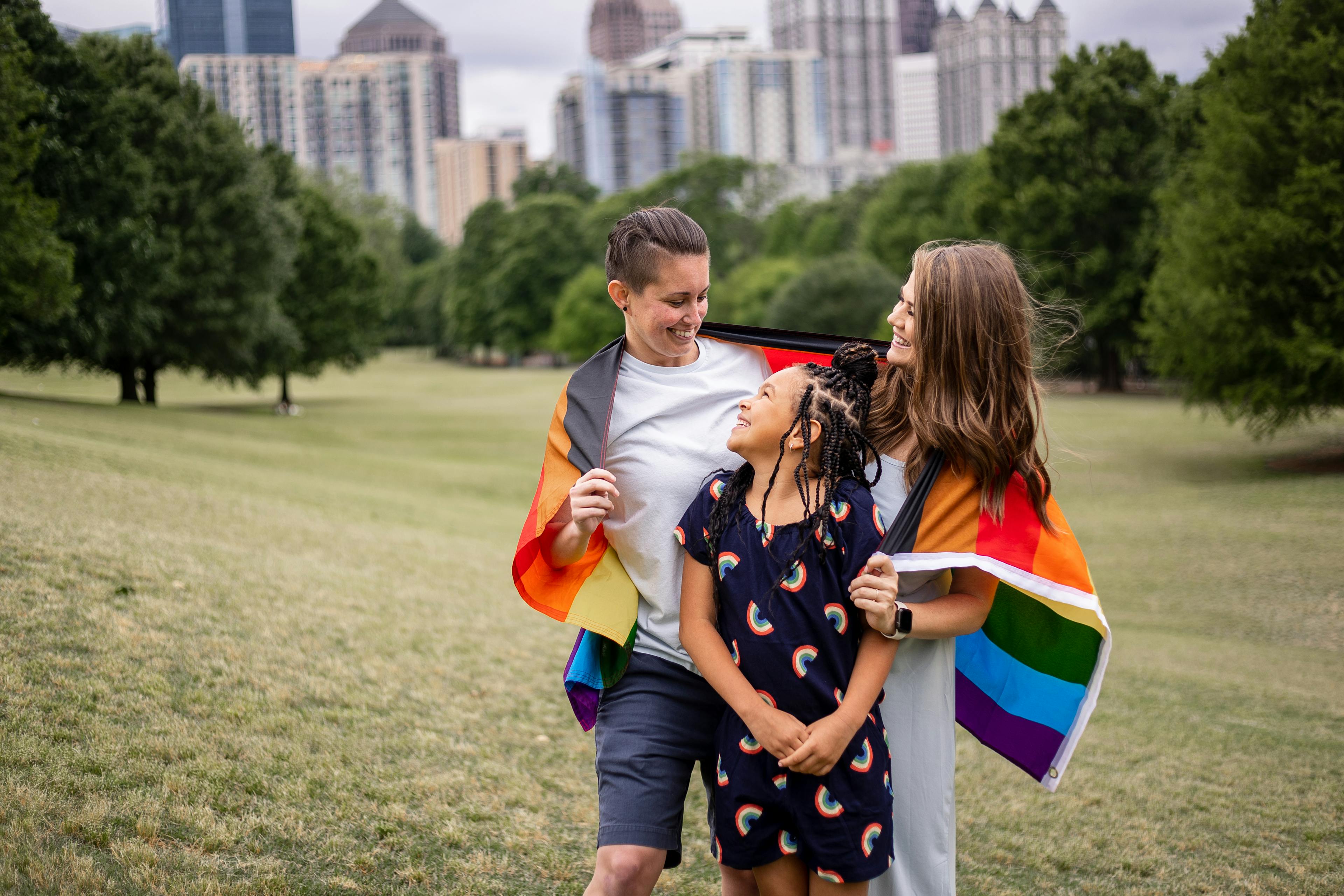 Three people smiling and holding a rainbow flag in a park with city buildings in the background.