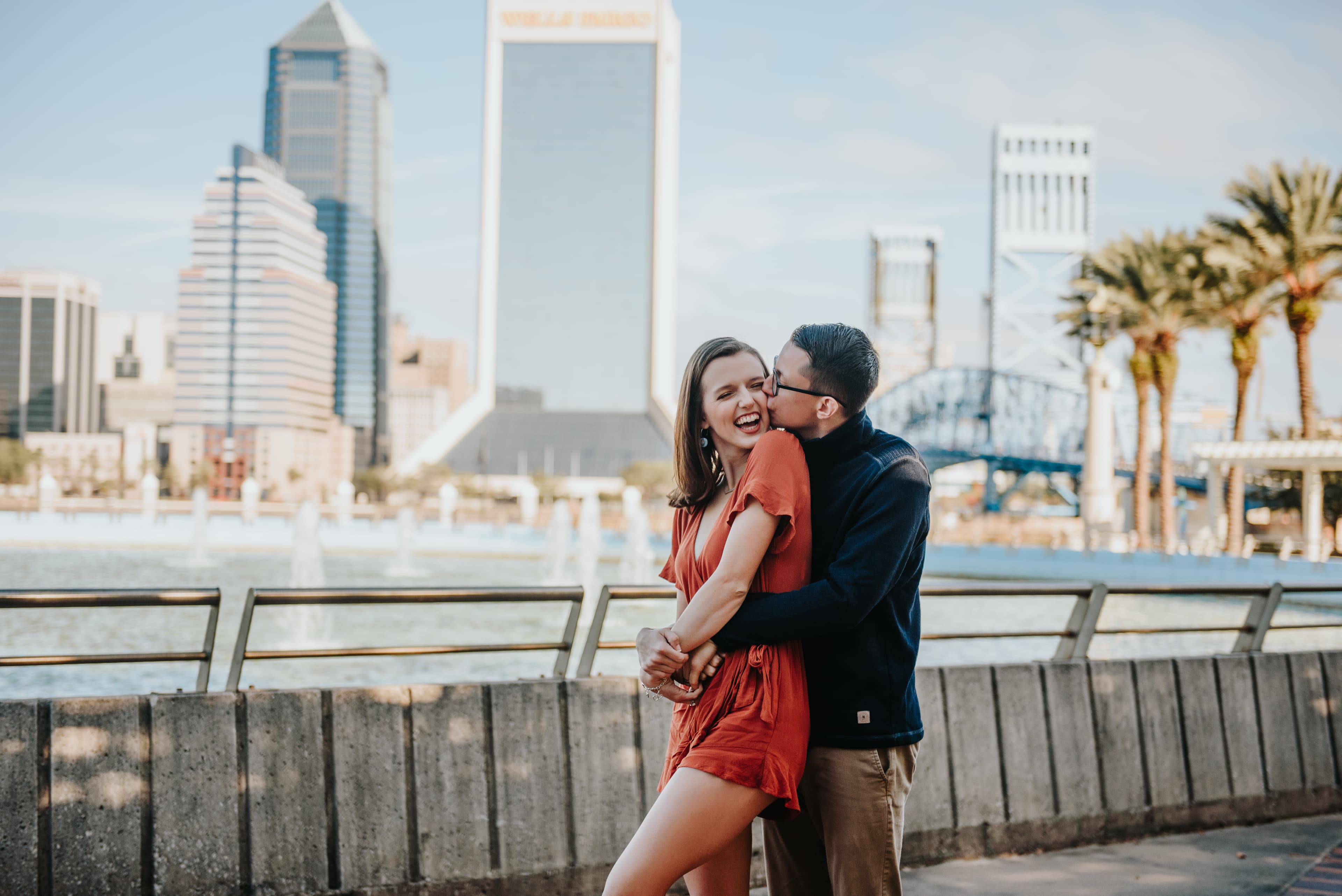 A joyful couple embracing and laughing by the waterfront with a city skyline in the background.