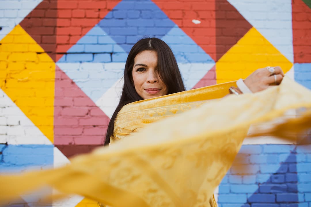 A woman holding a yellow scarf with a colorful geometric mural in the background.