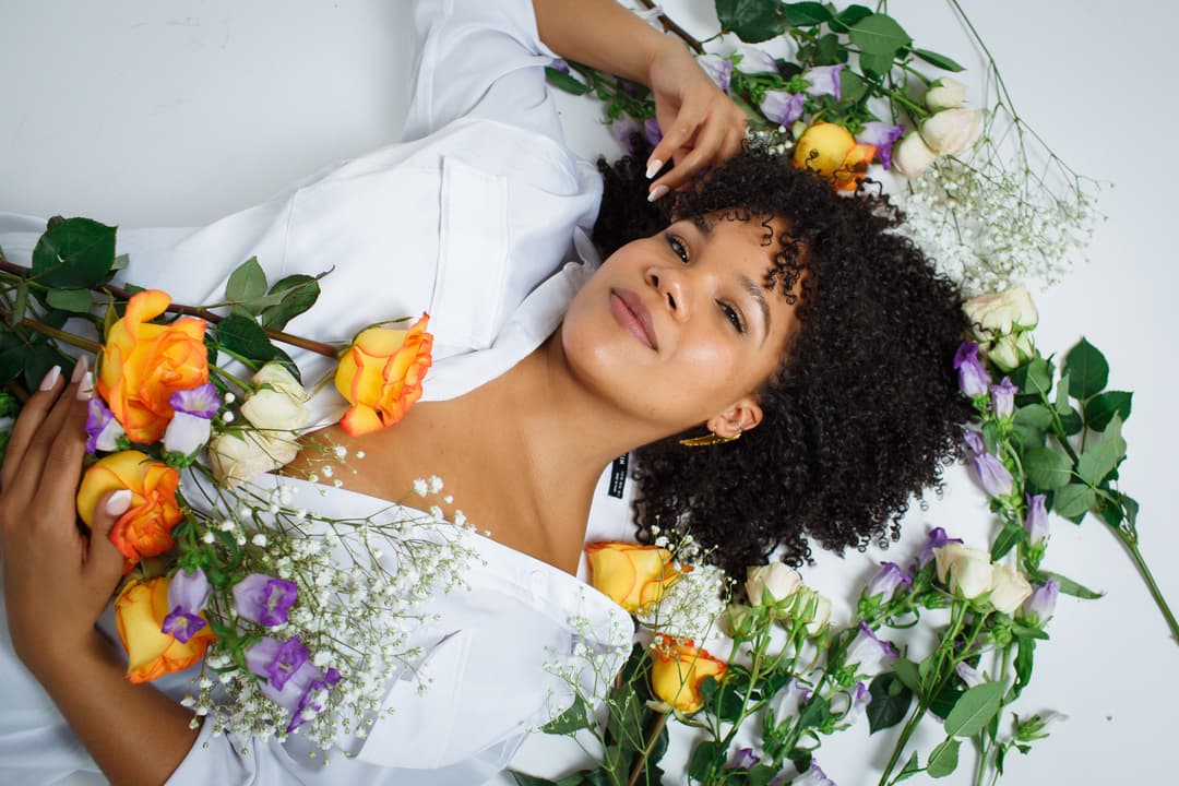 A woman lying surrounded by colorful flowers, looking up at the camera with a gentle smile.