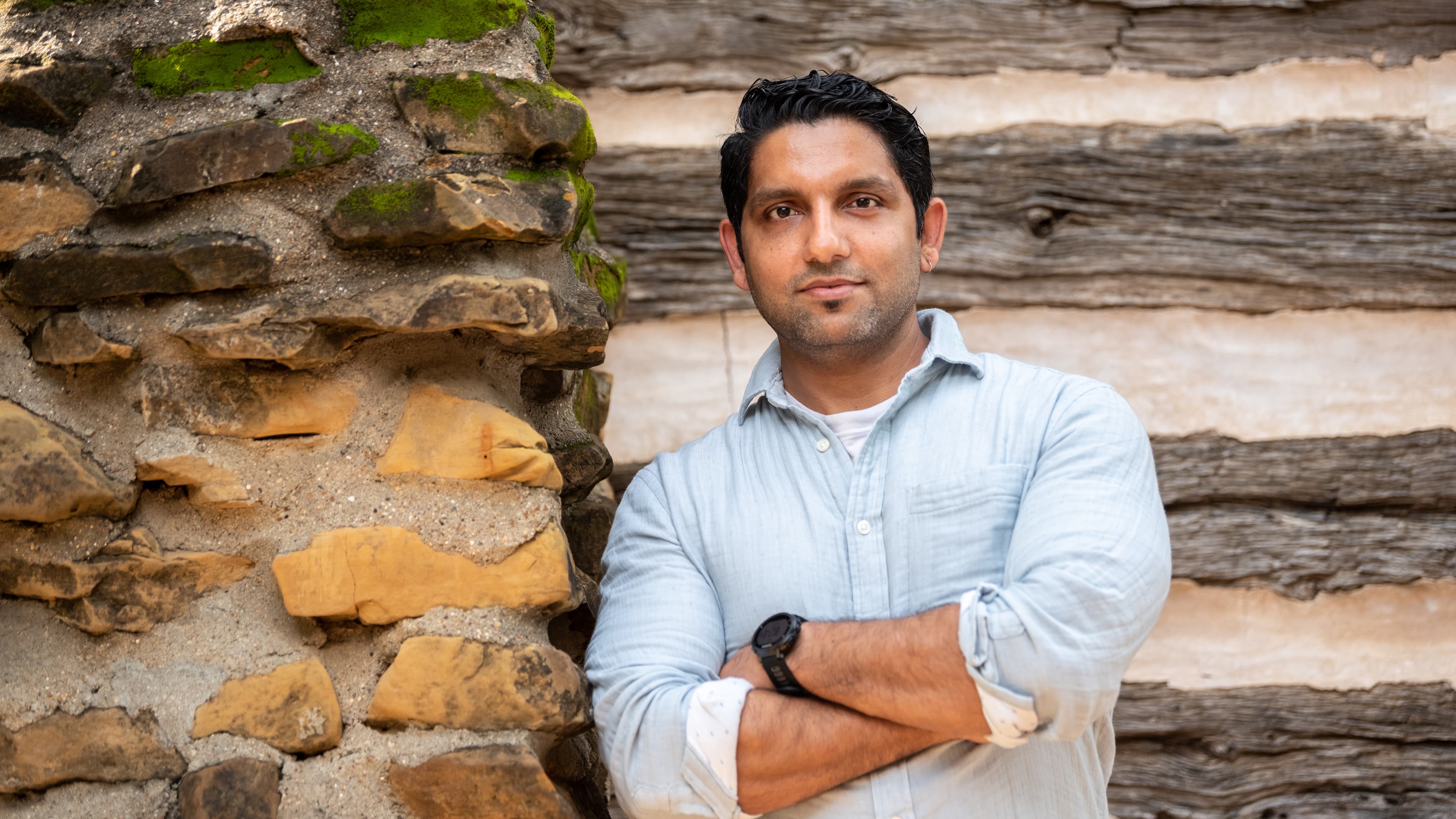 A man standing with arms crossed in front of a textured stone and wood wall.