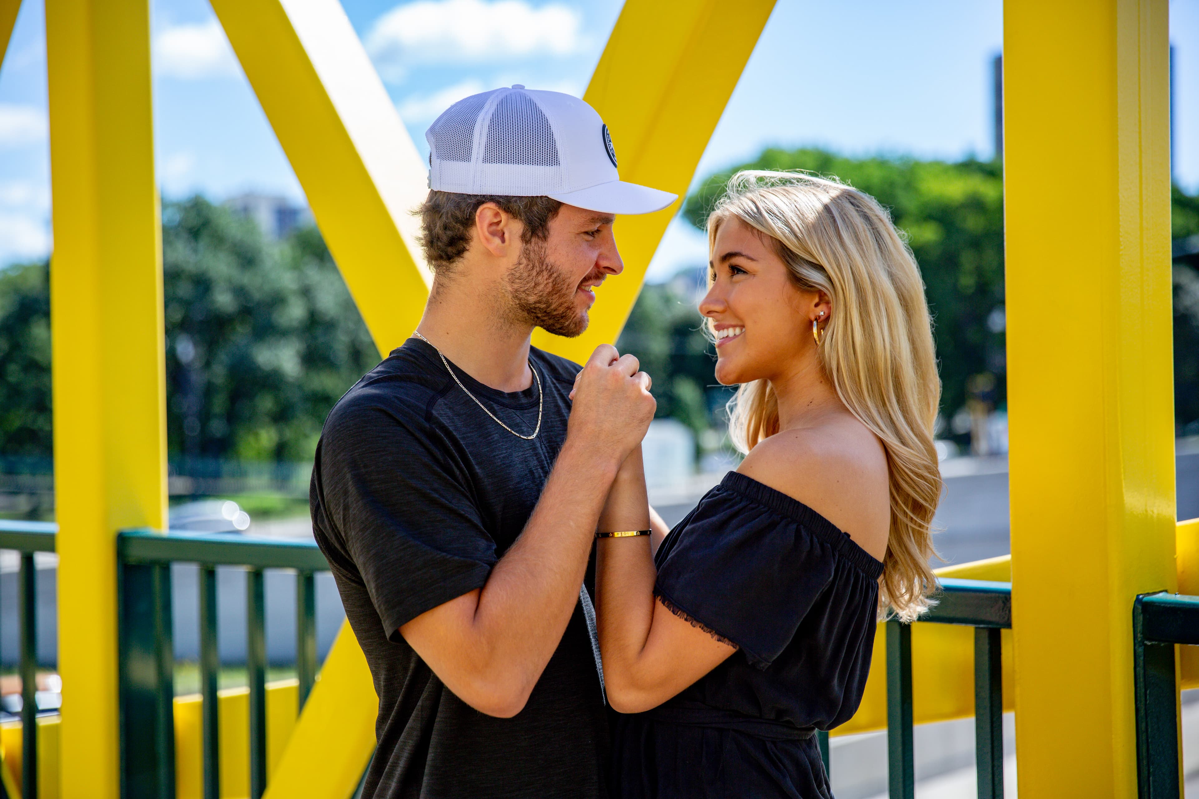 A smiling couple holding hands and looking at each other on a bridge with bright yellow beams.