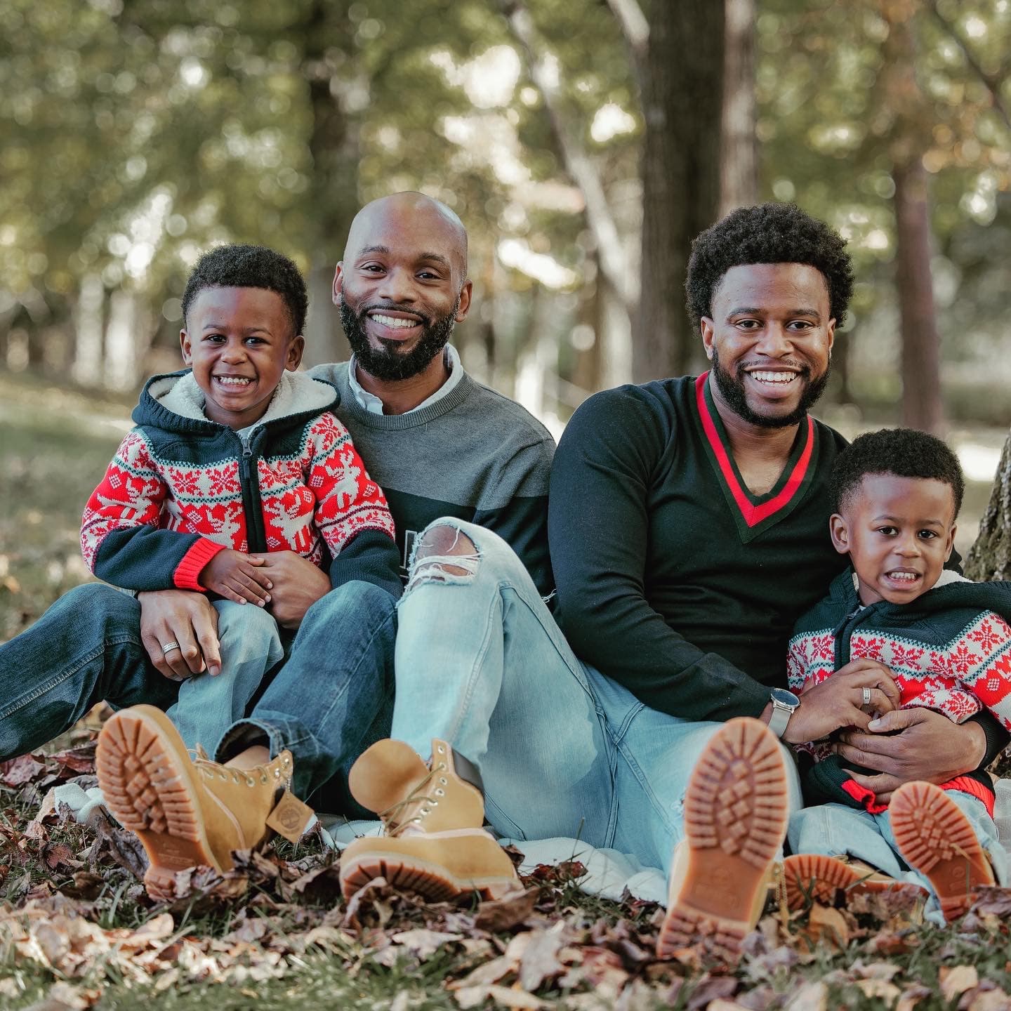 A happy family with two men and two young boys wearing matching sweaters, smiling and sitting together outdoors among fallen leaves.