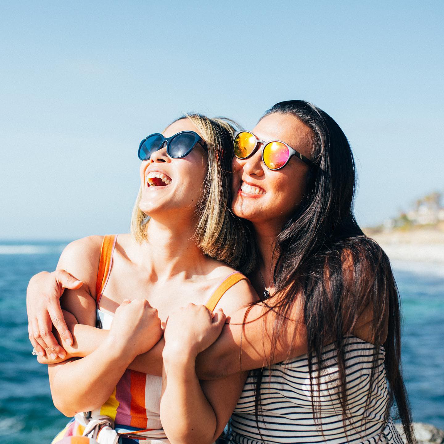 Two friends hugging and laughing by the sea wearing sunglasses.